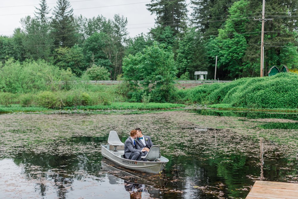  Two grooms share a kiss sitting in a small boat on a lake. 