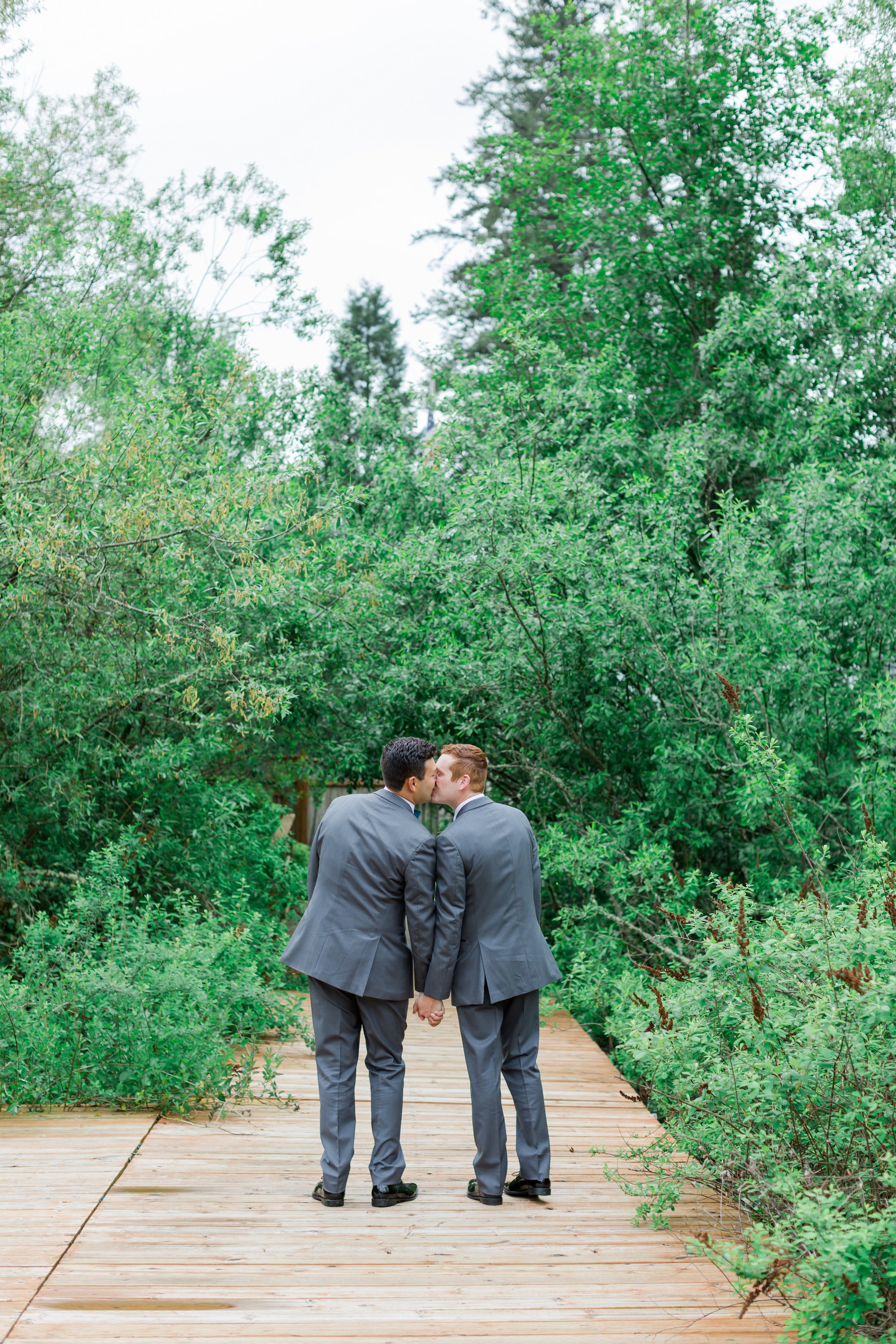  Two grooms share a kiss facing away from the camera in front of a lush green backdrop. 