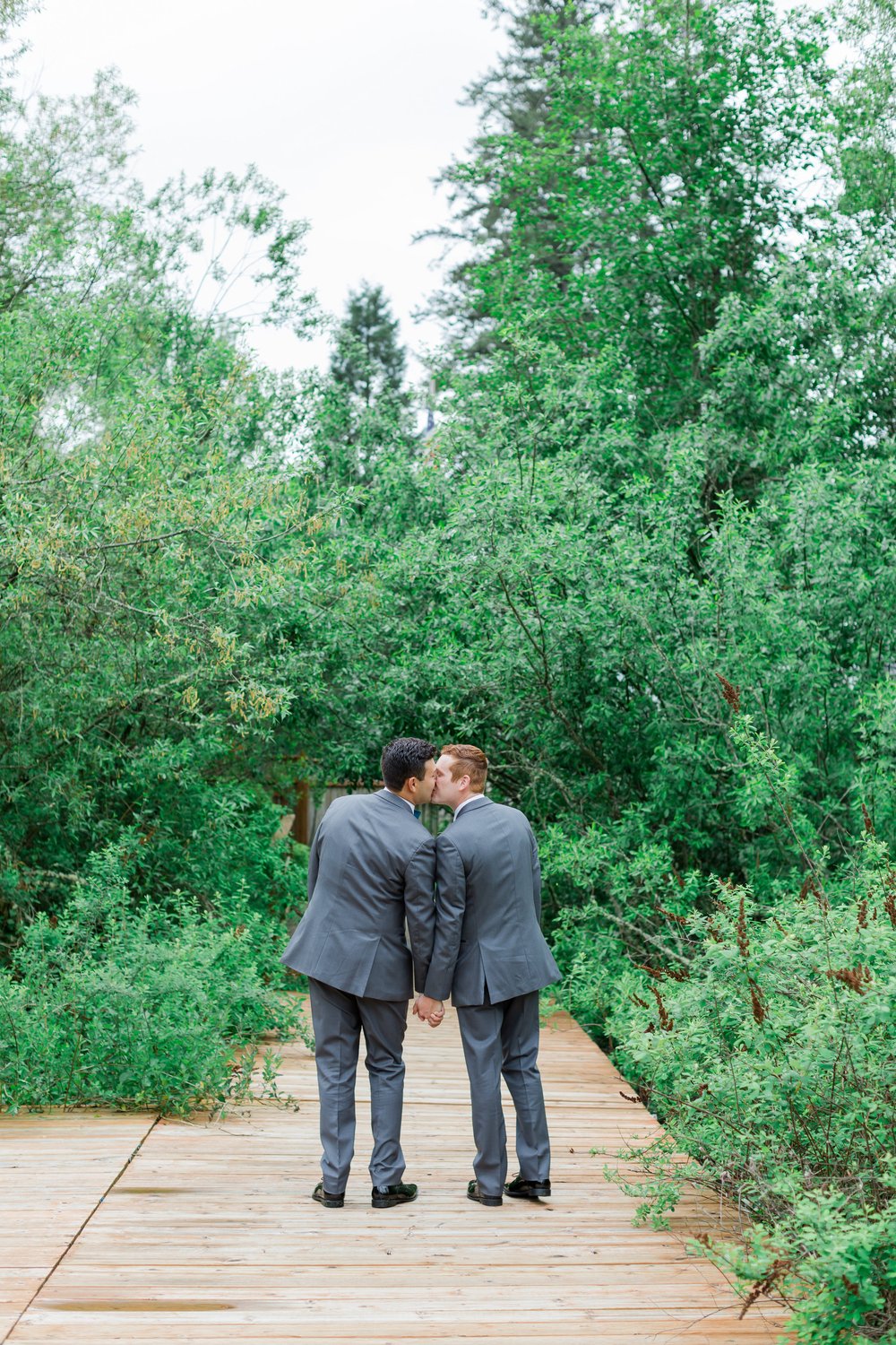  Two grooms share a kiss facing away from the camera in front of a lush green backdrop. 