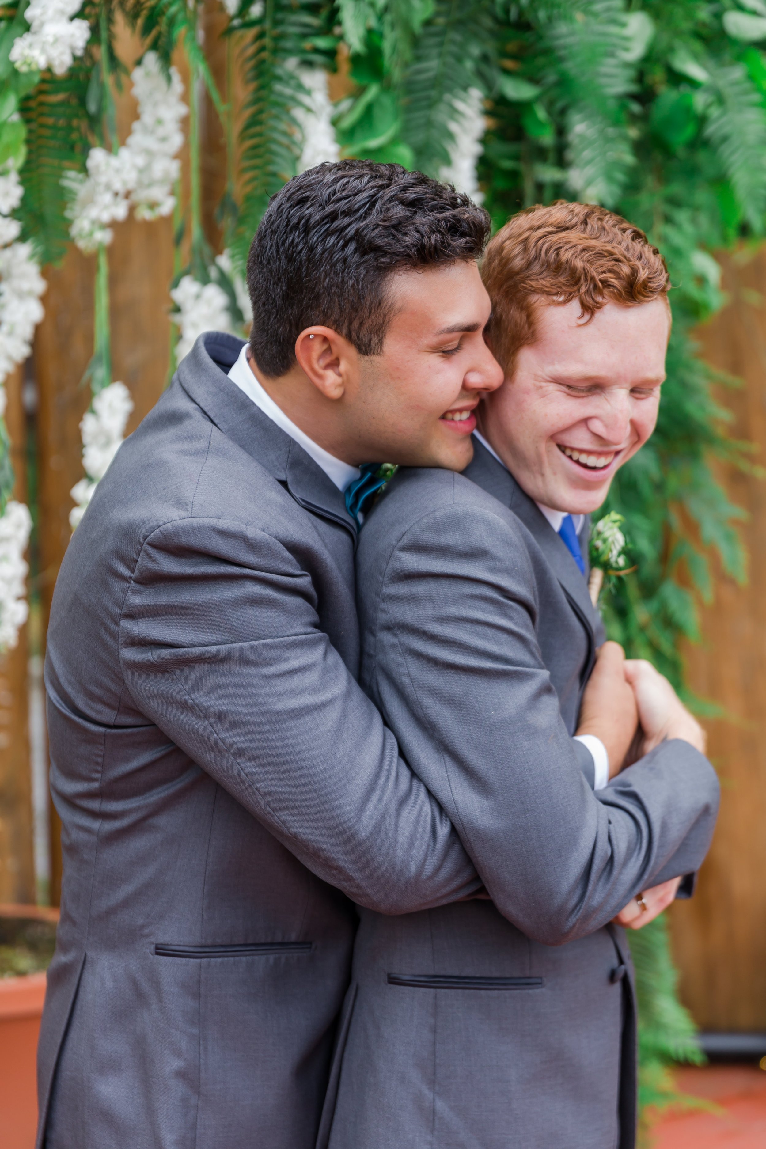  Two grooms embrace. There are green and white florals in the background. 
