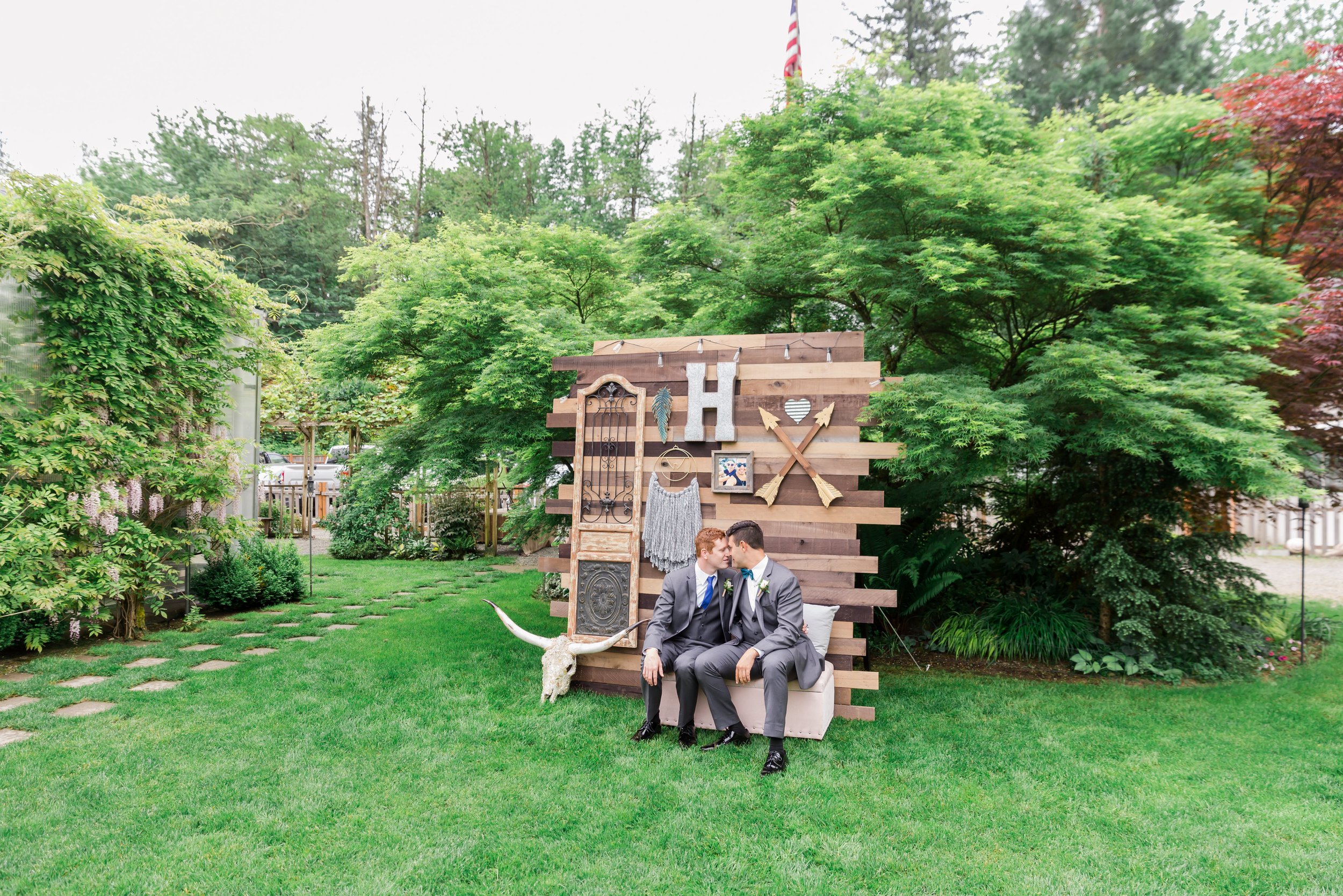  Two grooms sit in front of a photo backdrop with lush green trees in the background. 