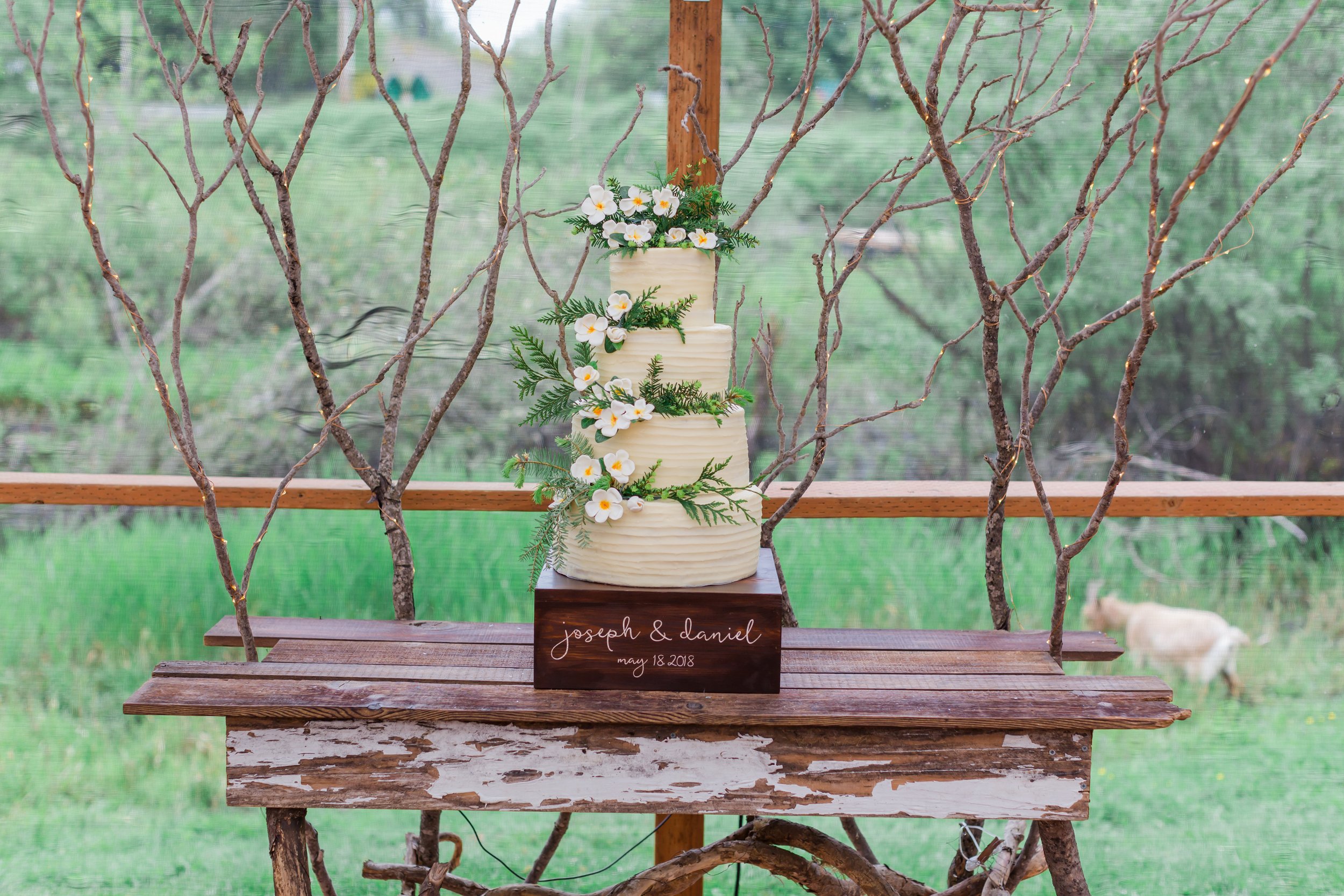  A wedding cake decorated with greenery and white flowers sits atop a cake stand that reads “Joseph &amp; Daniel, May 18, 2018” 
