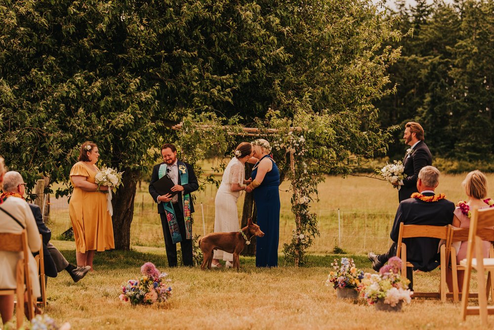  Two brides hold hands during their wedding ceremony. Their dog is standing with them at the altar. 