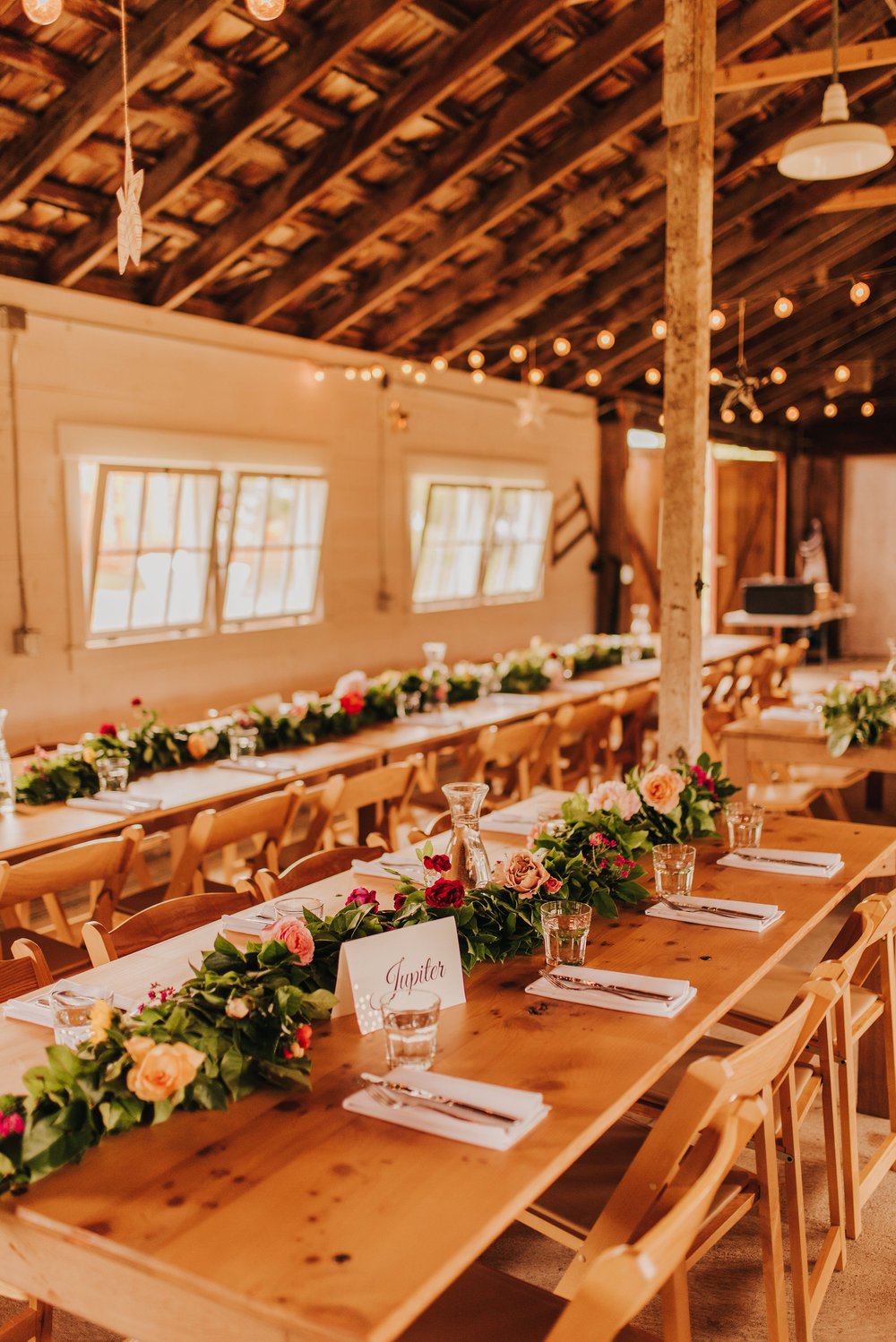  Tables are set in a barn for a wedding reception. There are floral runners down the middle of the tables and cafe lights hanging from the ceiling. 