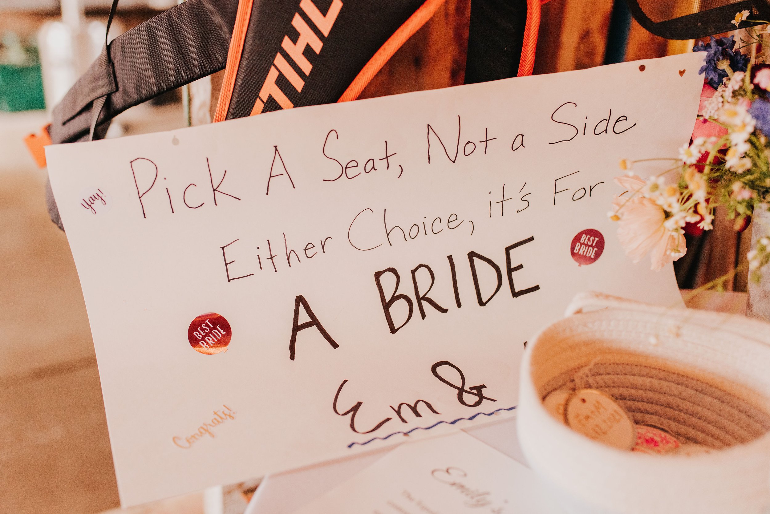  A sign on white paper reads “Pick A Seat, Not A Side, Either Choice, it’s For a  Bride” 