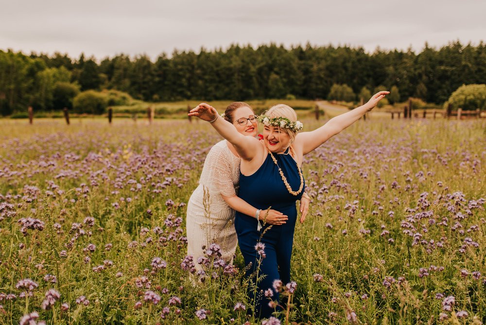  Two brides pose in a meadow of purple wild flowers. One bride has her arms outstretched. 