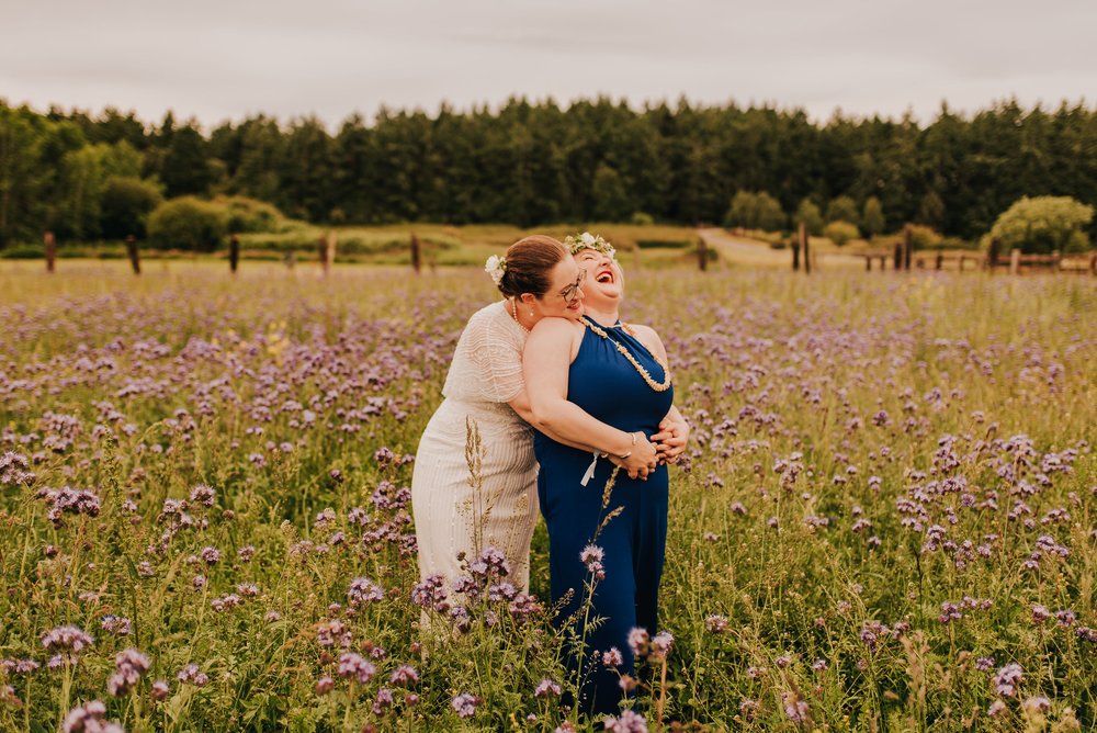  Two brides pose in a meadow of purple wildflowers. One bride is laughing while being hugged from behind by the other bride. 