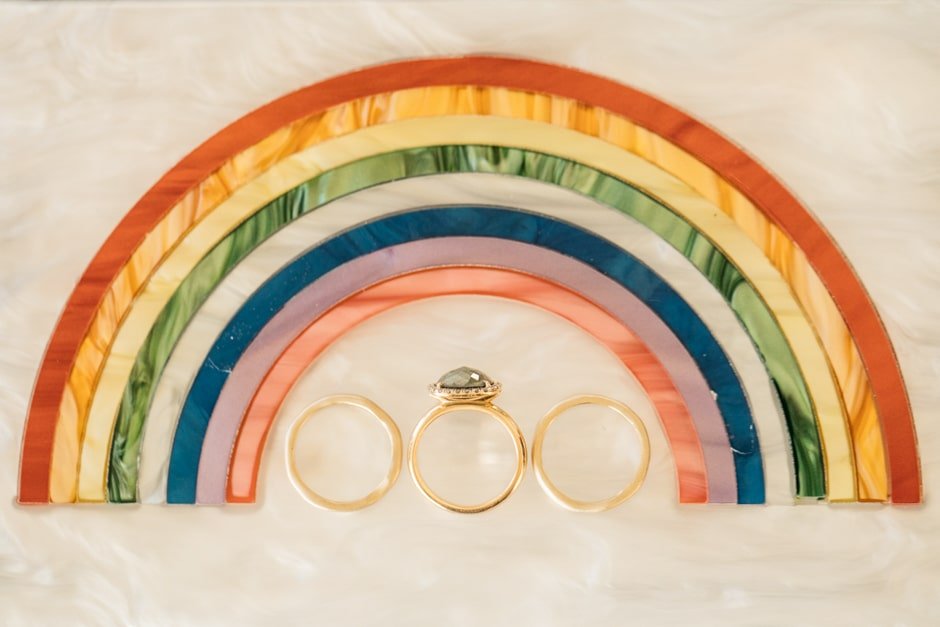  Three wedding rings are placed on a flat surface that with a rainbow above them. 