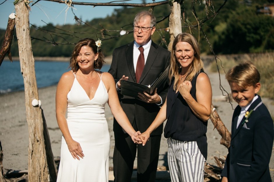  Two brides are standing at an altar made of driftwood with their officiant and children. One bride is making a celebratory fist pump with her right hand. 