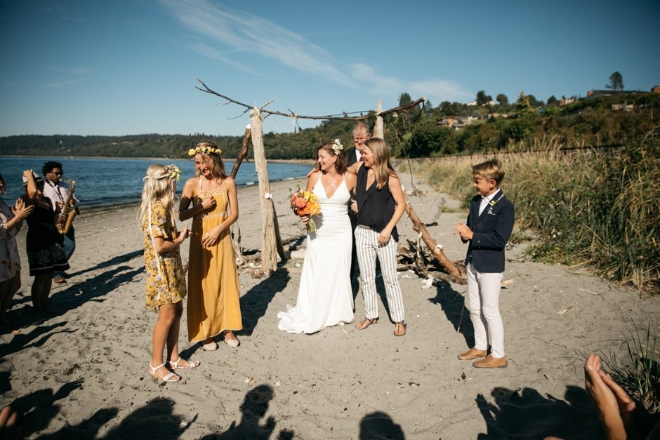  Two brides stand at a driftwood altar with their officiant and children during their wedding ceremony. 