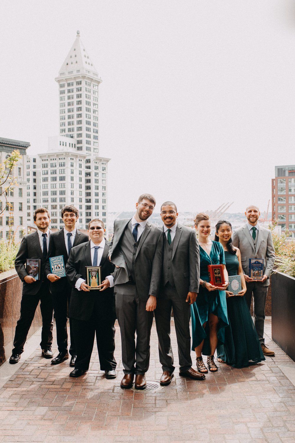  Two grooms and their wedding party pose on a brick sidewalk with Smith Tower in the background. 