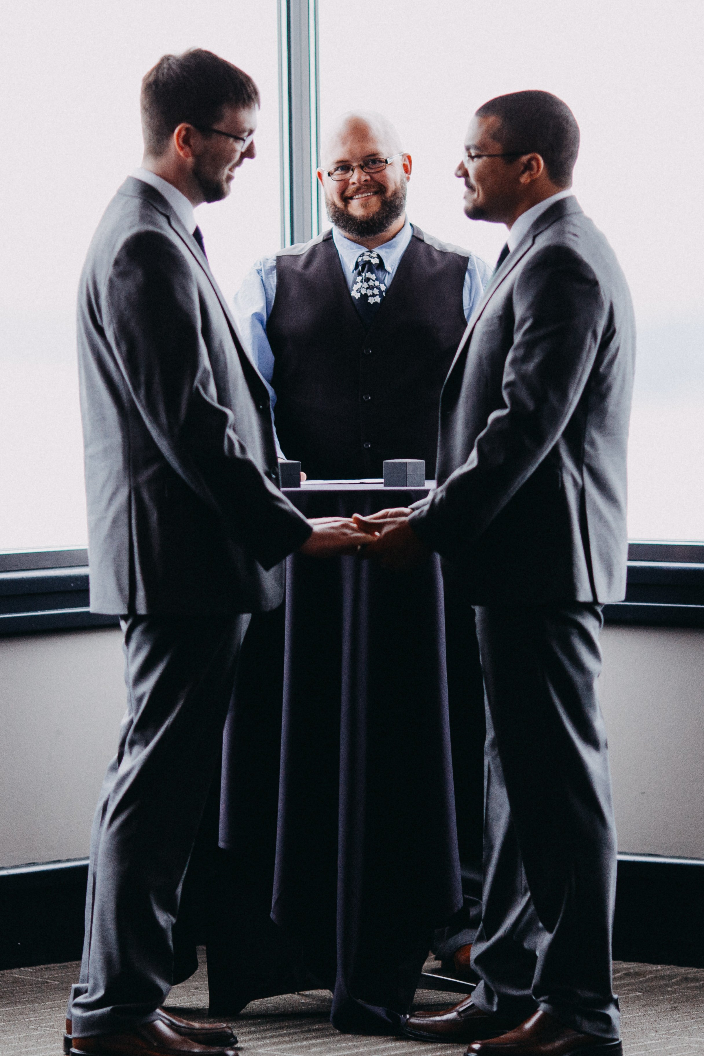  Two grooms hold hands during their wedding ceremony. Their officiant smiles behind them. 