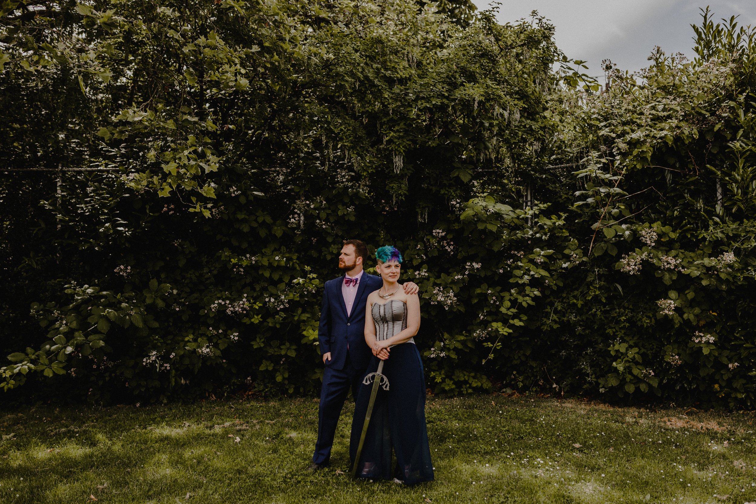  A groom and his future spouse are posed together in front of some lush greenery. The person on the right holds a sword pointed toward the ground. 