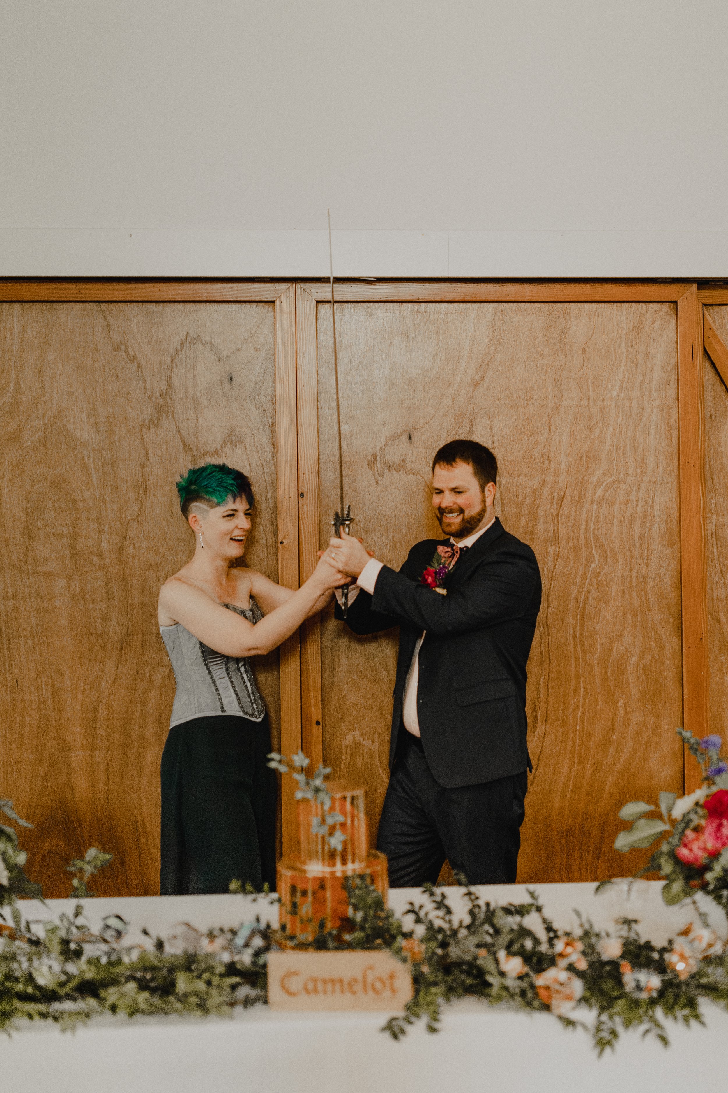  A marrier and their groom hold a sword in the air between them above their wedding cake in preparation of cutting the cake with the sword. 