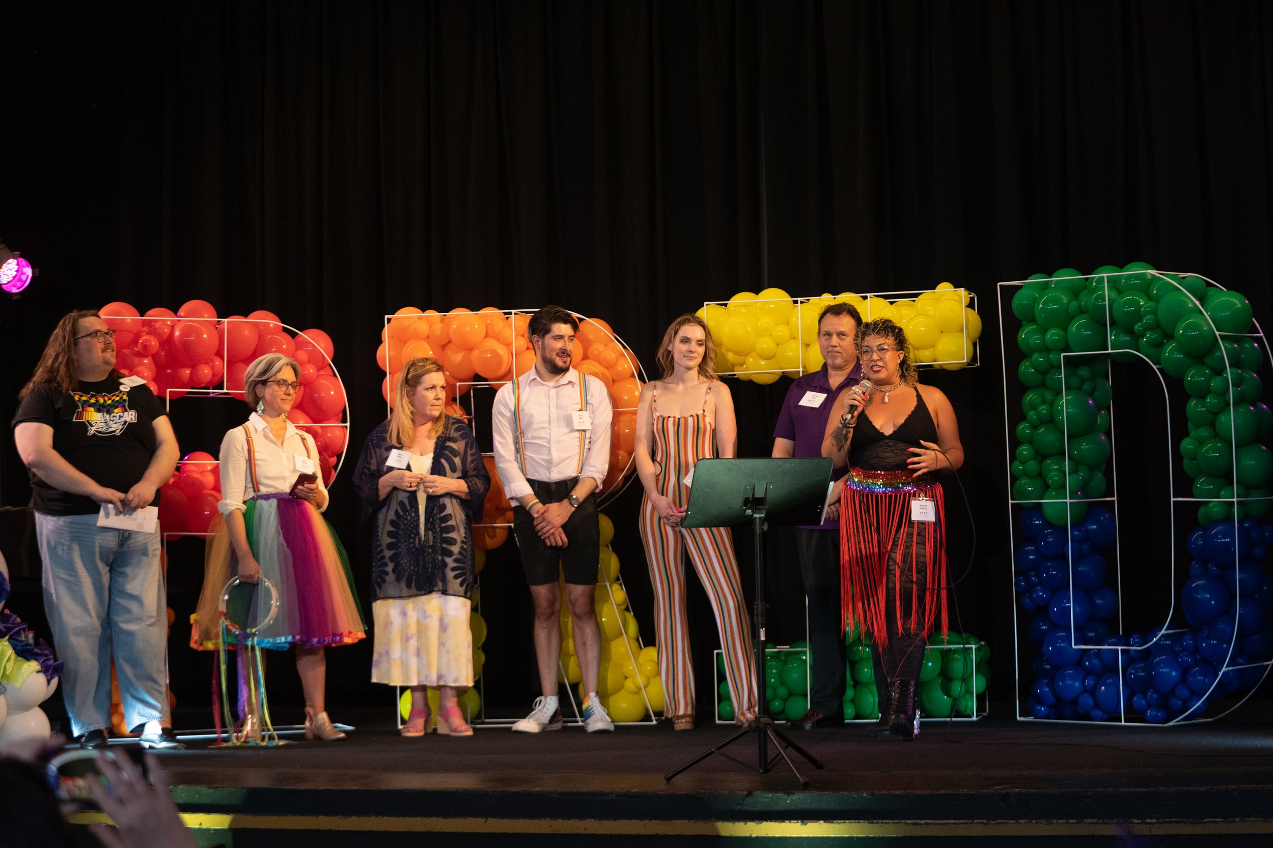  A group of event professionals stands on a stage in a line. There is a display behind them of the word “pride” in rainbow balloons. 
