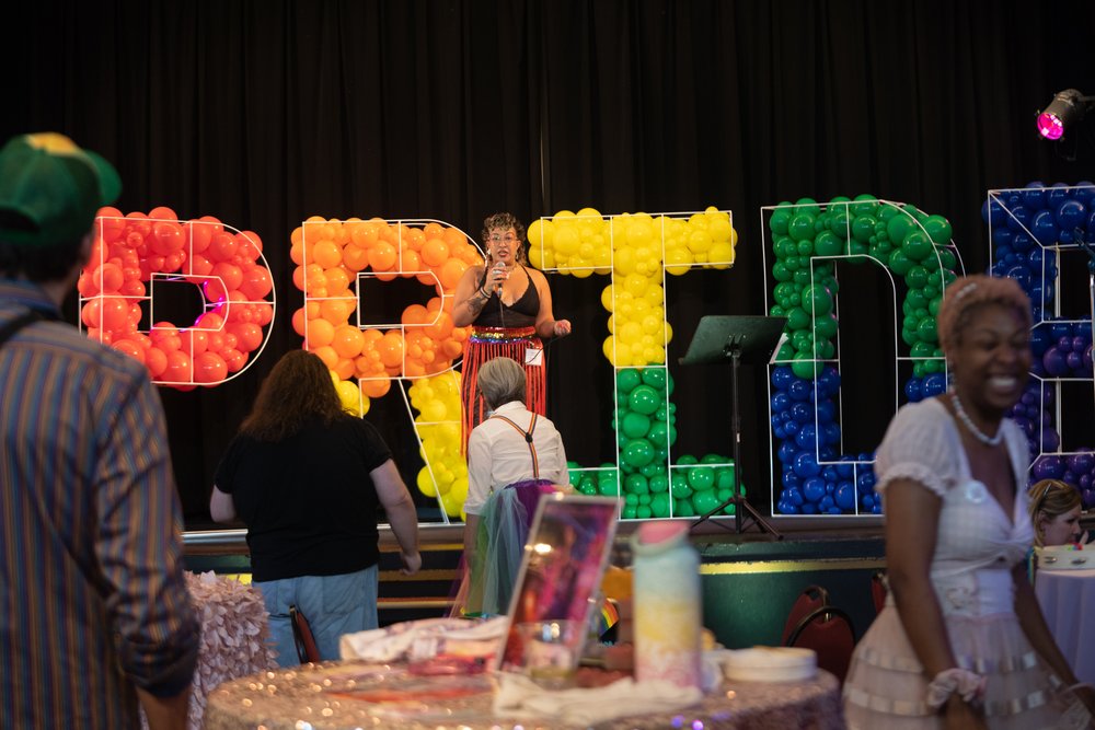  The event hostess is on the stage in front of a display that reads “pride” in rainbow balloons. 