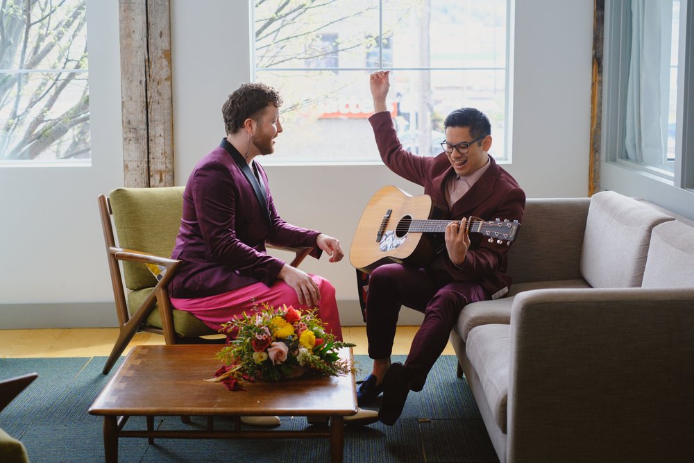 Two grooms sit across from each other and sing a song. One is playing the guitar. 