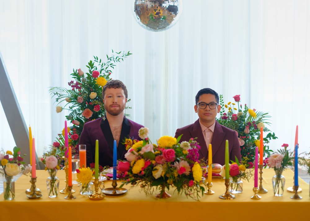  Two grooms look into the camera, sitting at a table draped in mustard yellow and adorned with colorful florals and candles. 
