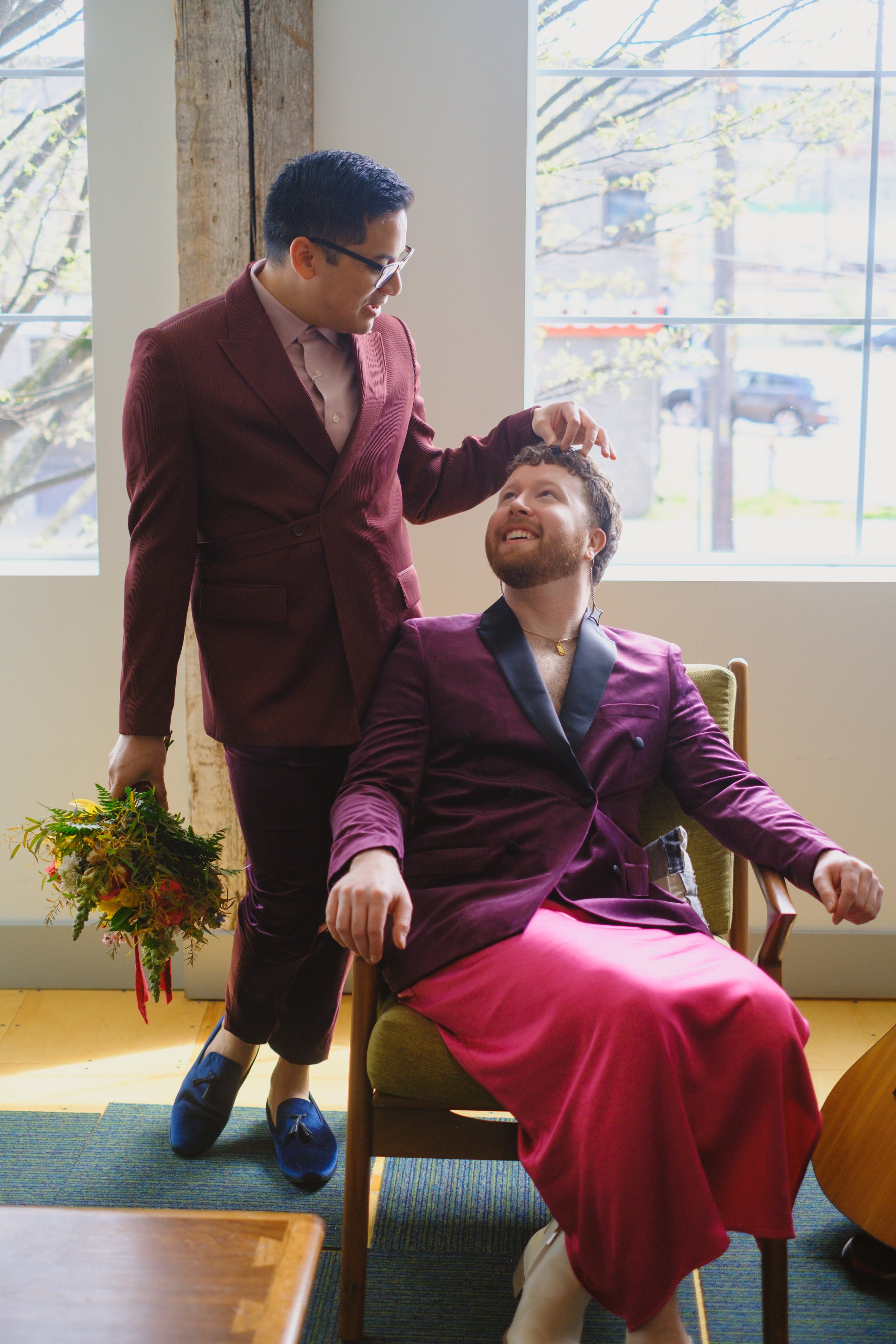  A groom stands next to his future husband who is seated. He is playing with his hair with one hand and holding a colorful bouquet with the other. 