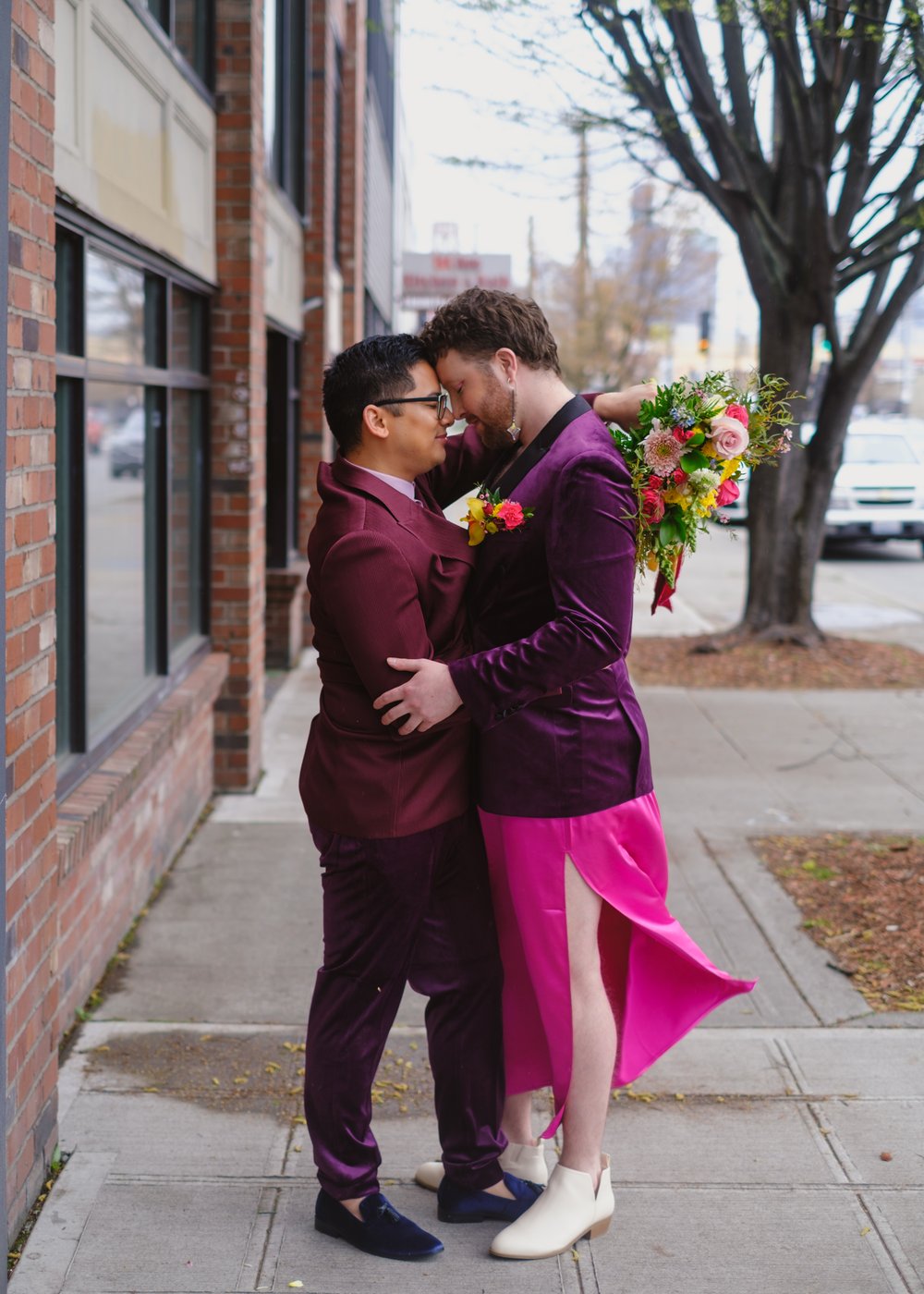  Two grooms embrace on the sidewalk in front of a brick building. One is wearing a pink skirt that is gently billowing behind them in the wind. 