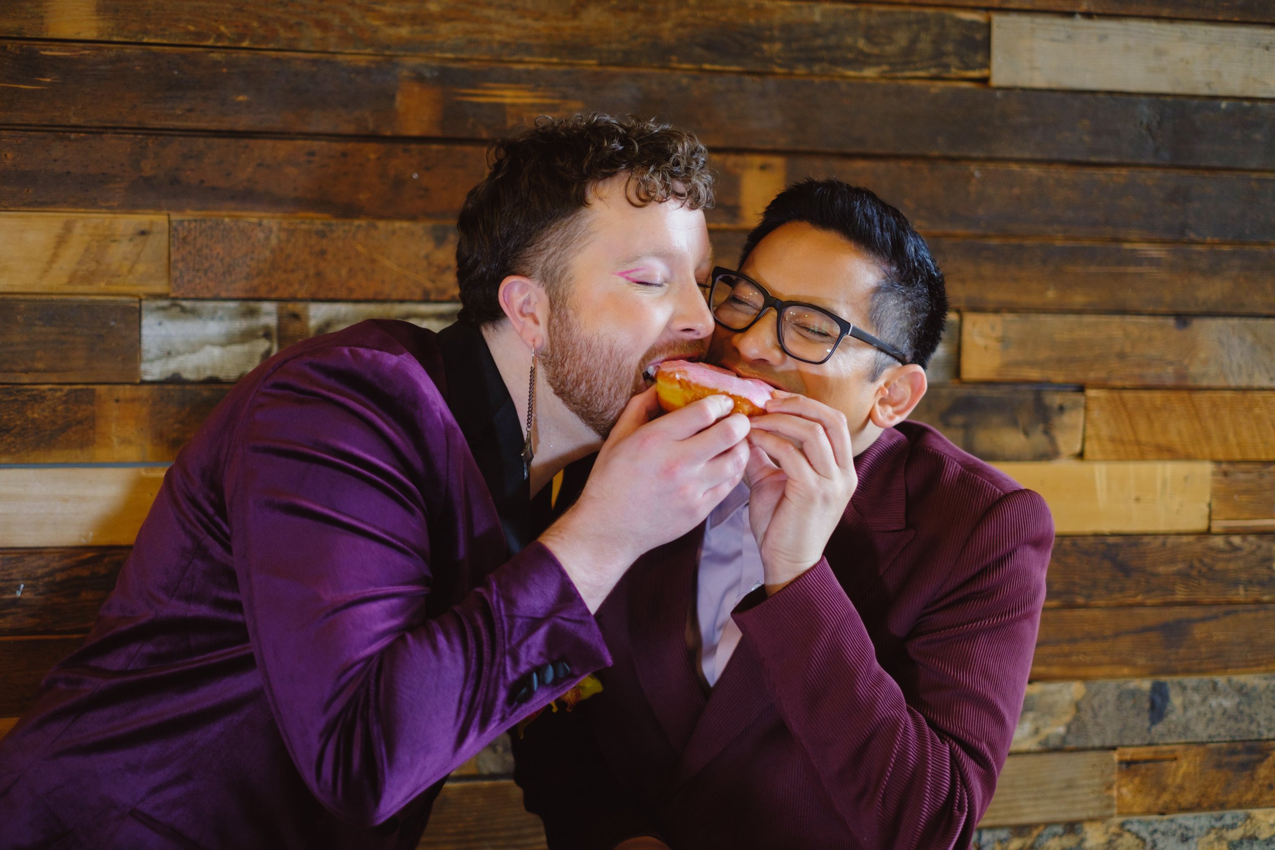  Two grooms both take a bite of the same donut. 