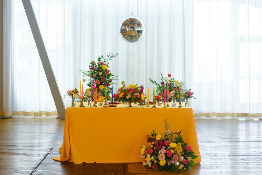  A table with a mustard yellow linen, colorful floral arrangements, and mismatch taper candles in brass holders. A disco ball hangs above and behind. 