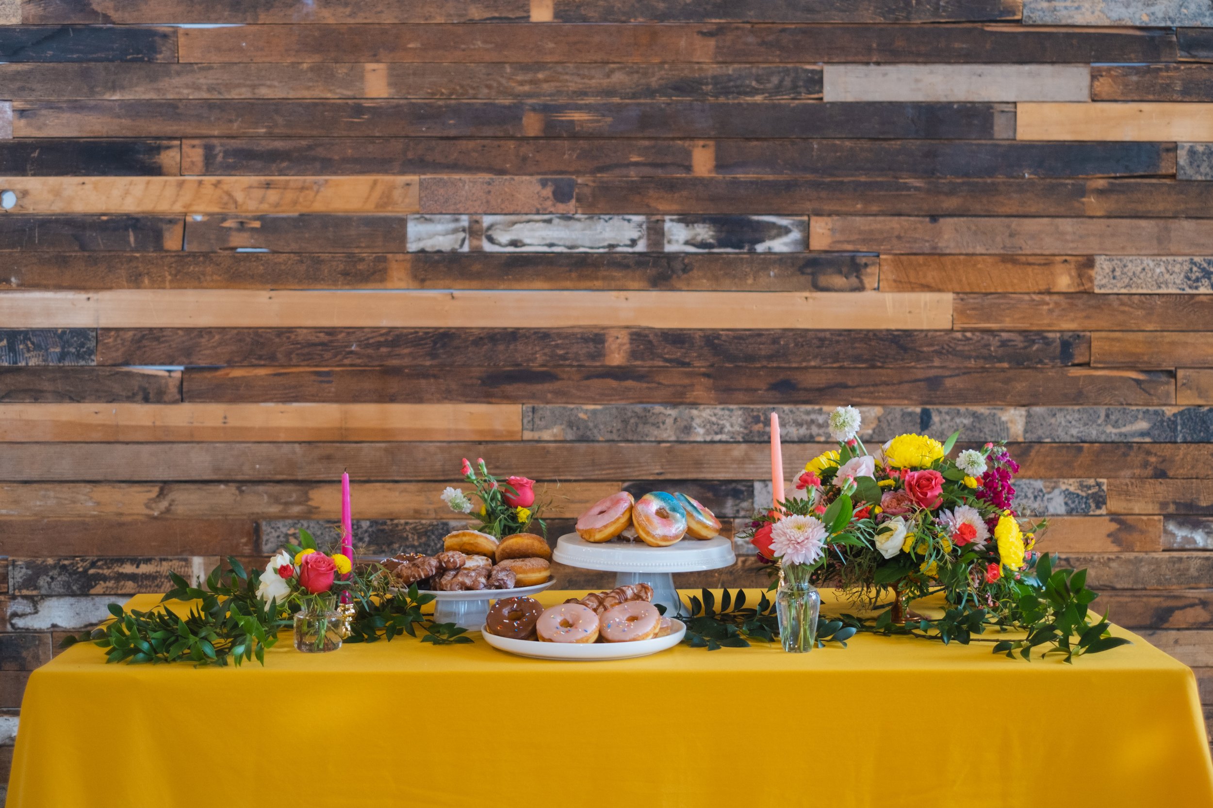  A display of different types of donuts on cake plates in front of a wood paneled wall. 