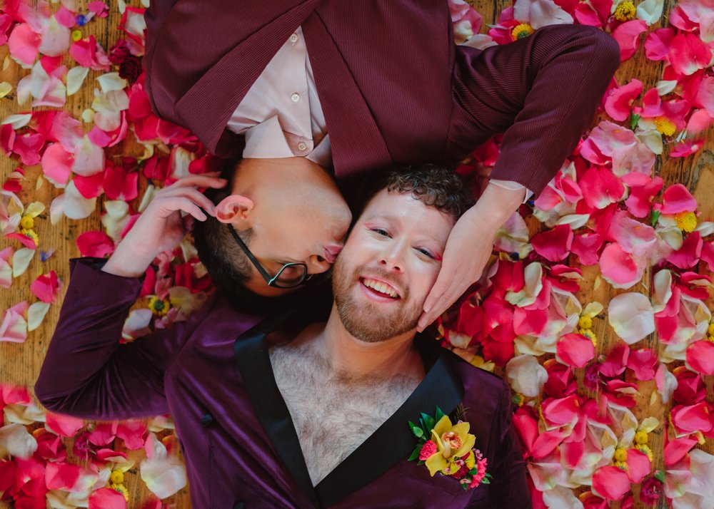 Two grooms lay in a bed of pink, white, and yellow flower petals. One kisses the other on the cheek. 