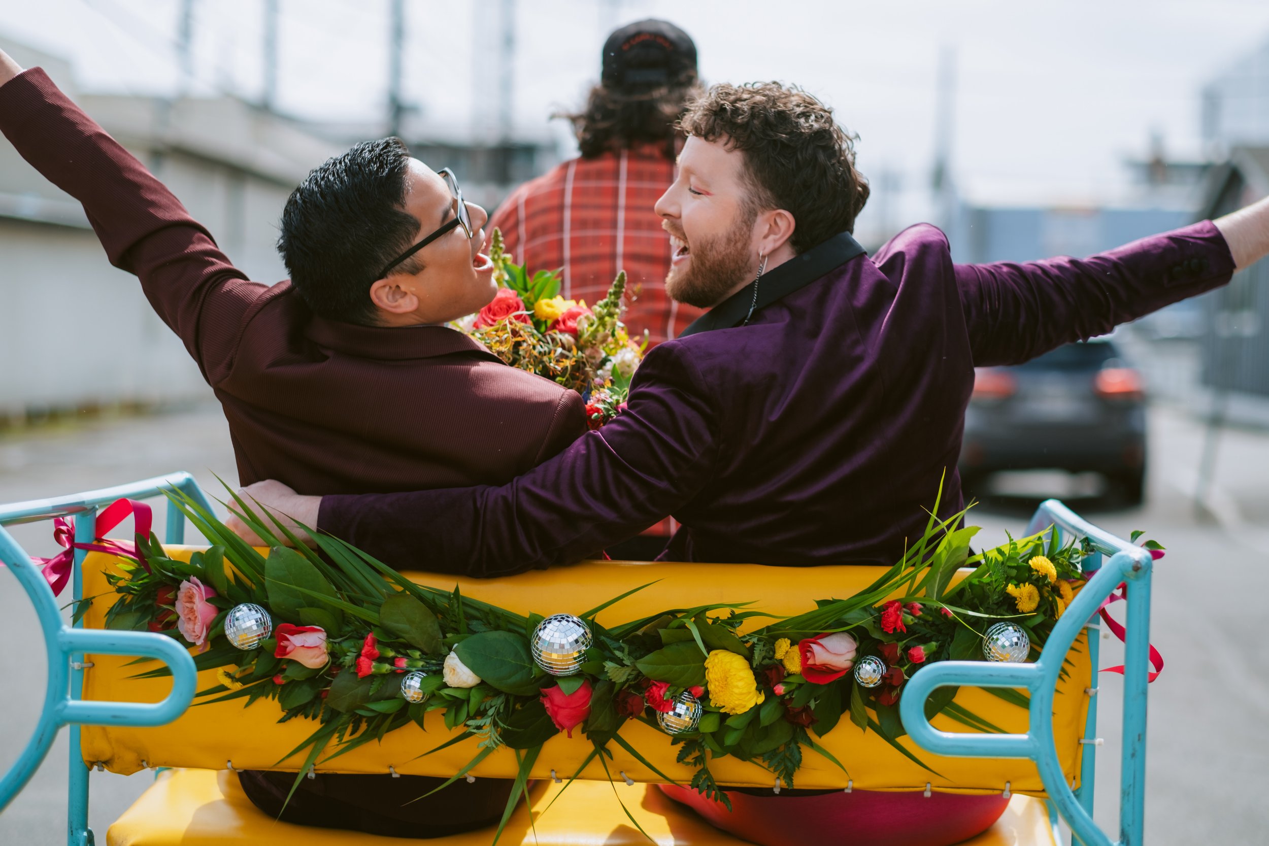  Two grooms ride in a pedi-cab with a swag of greenery, colorful flowers, and disco balls adorning the back of the cab. 