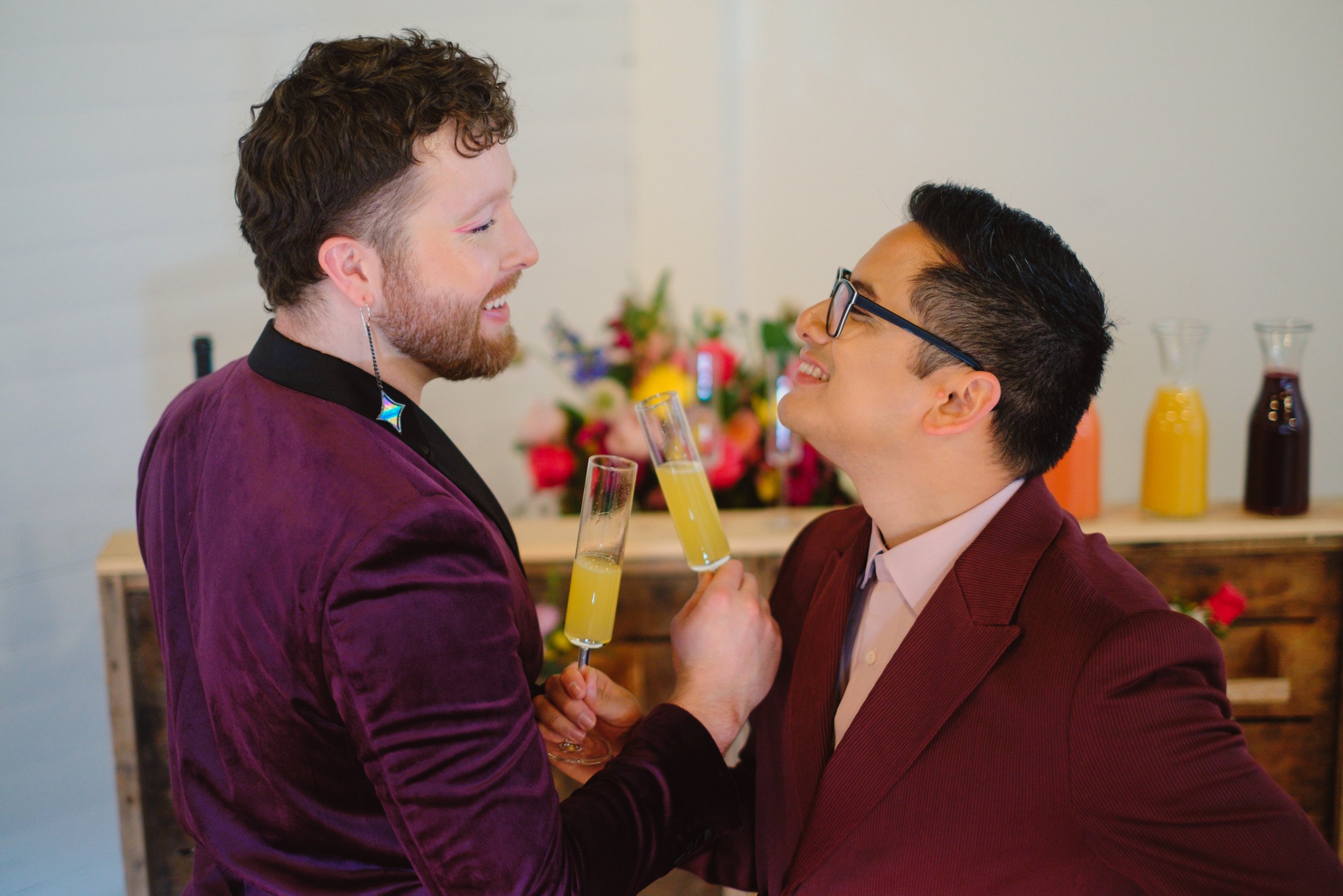  Two grooms smile at each other with their arms linked holding mimosas. 