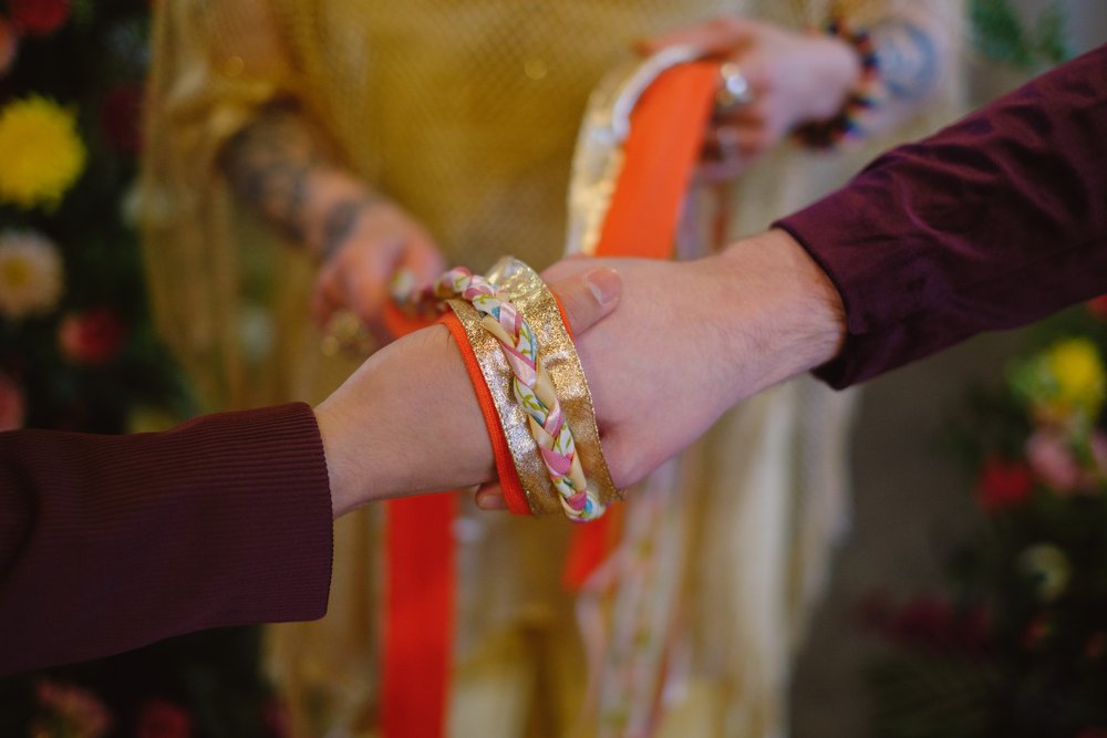  A close up image of two holding hands being wrapped with gold and orange braided ribbon. 