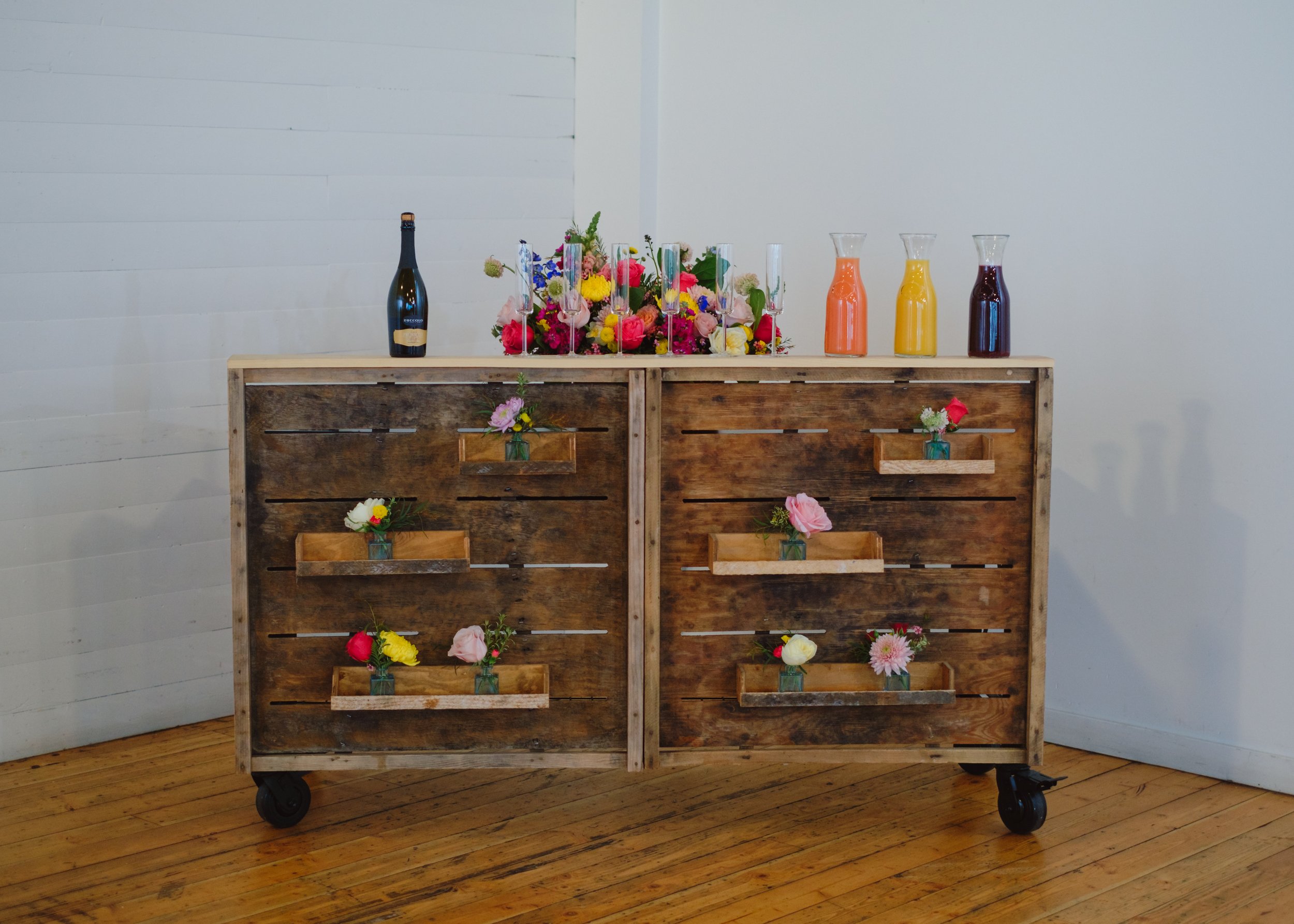  A wood slat style bar set with carafes of juice, champagne glasses, and a bottle of sparkling wine. 