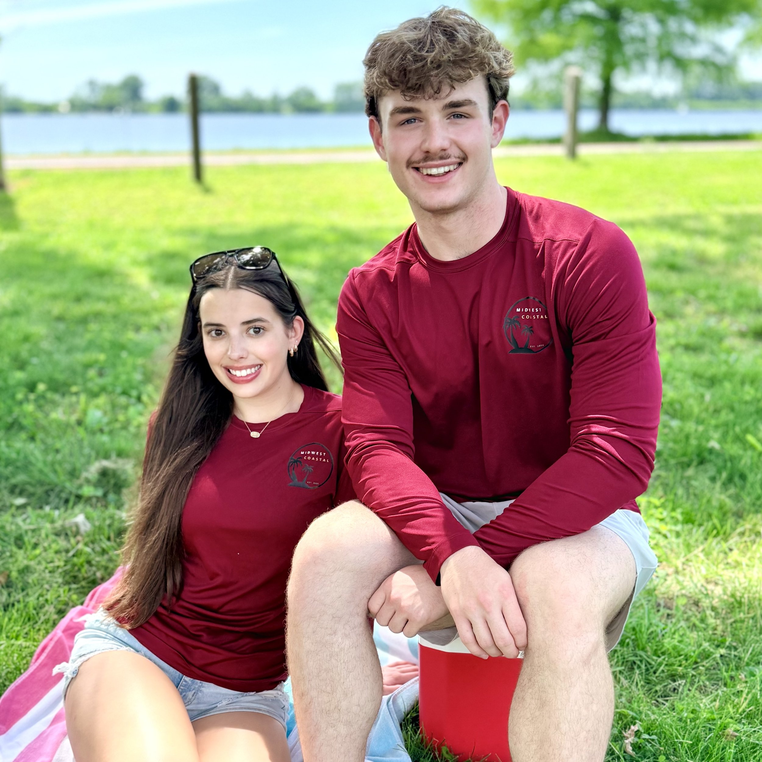 A young man with curly hair and a mustache wearing a red T-shirt with a palm tree logo, sitting outdoors on green grass near a body of water, holding a red cup, smiling while partially resting his arm on a woman's knee. The woman has long dark hair, wears sunglasses on her head, and has a friendly smile.