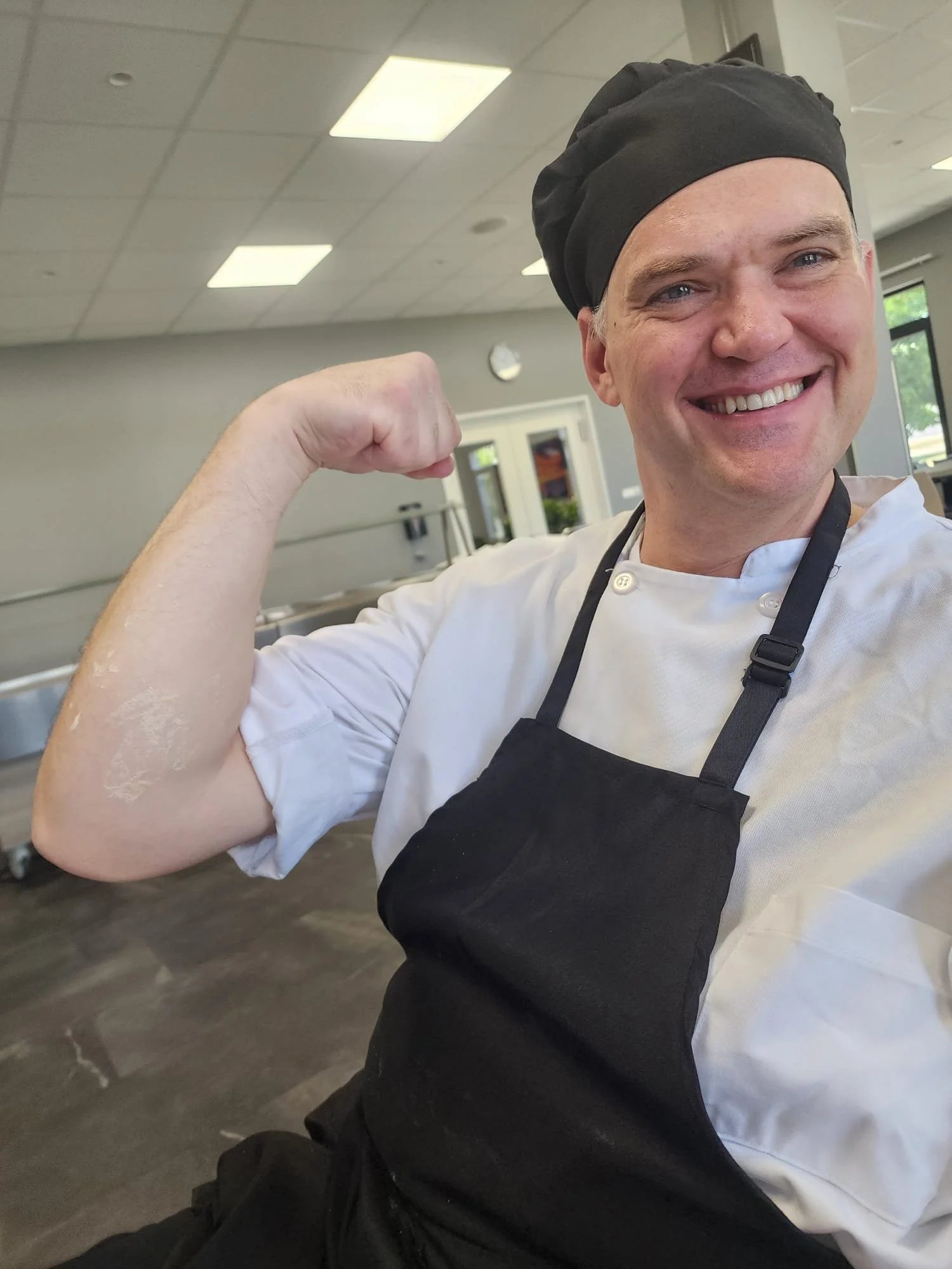 A smiling man dressed as a chef in a kitchen, flexing his right arm to show strength.