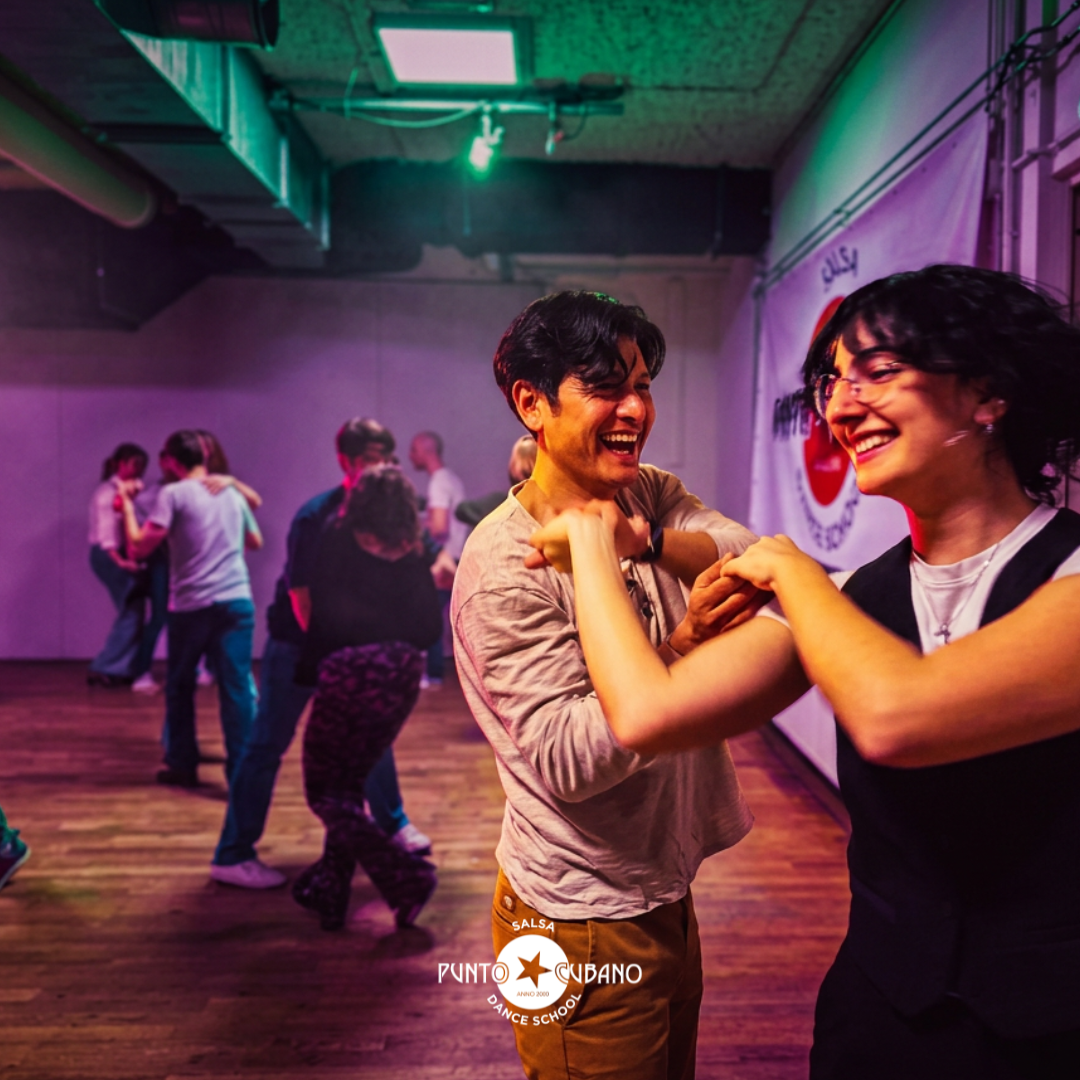 People in a dance studio holding hands and dancing in a circle, with a mirror and speakers in the background, promoting a salsa dance start course.