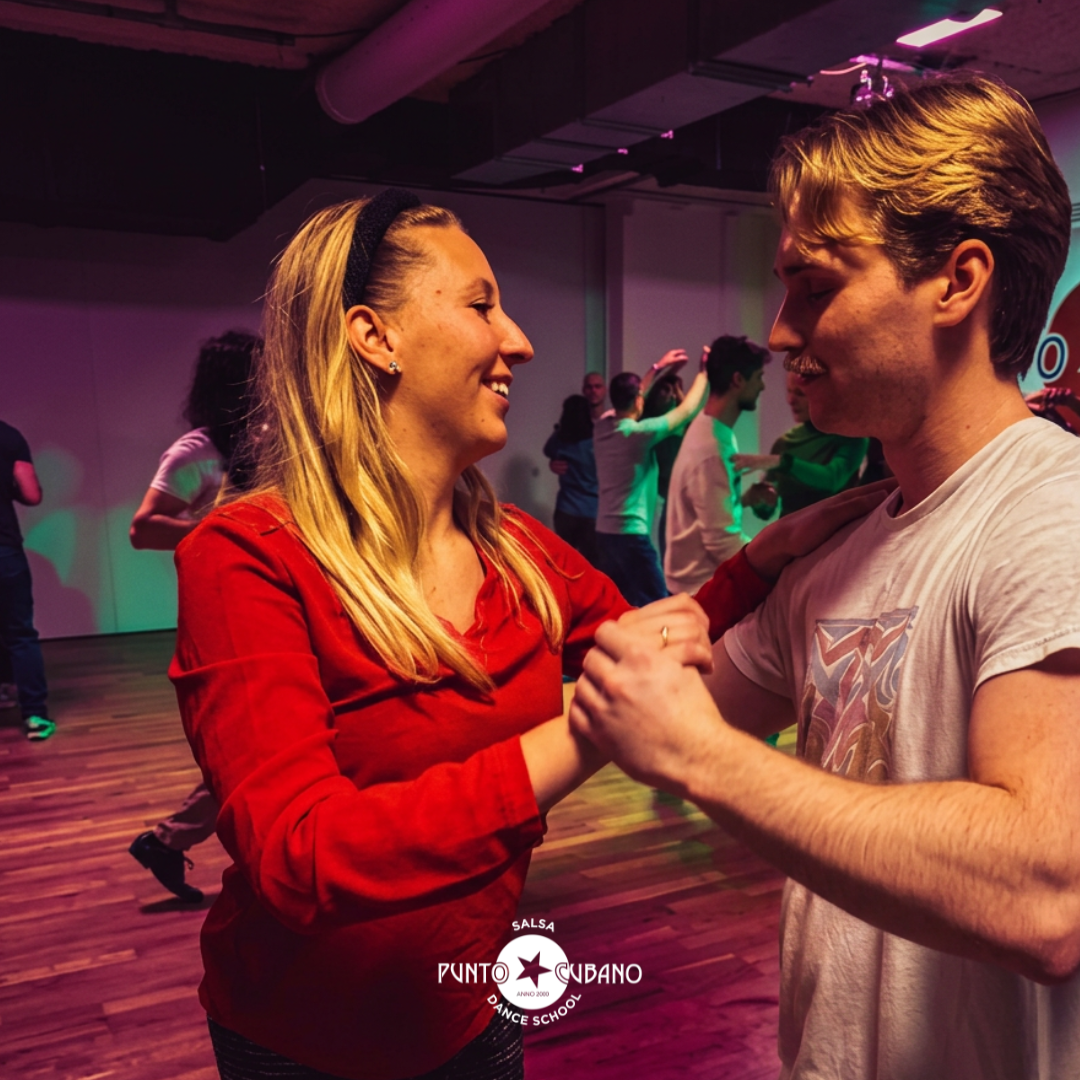 People dancing and practicing salsa steps in a dance studio with colorful lighting, socializing during a salsa dance class or event.