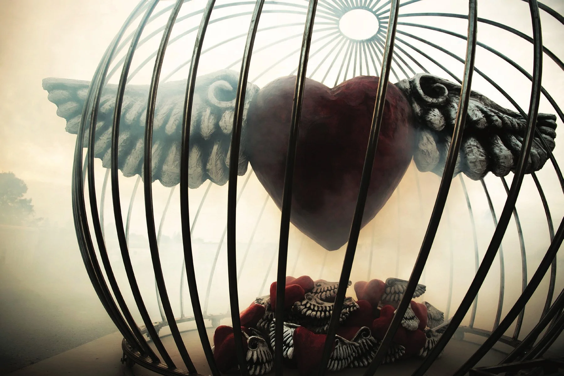 Image of a large winged heart inside a spherical cage, with a pile of smaller hearts beneath it.
