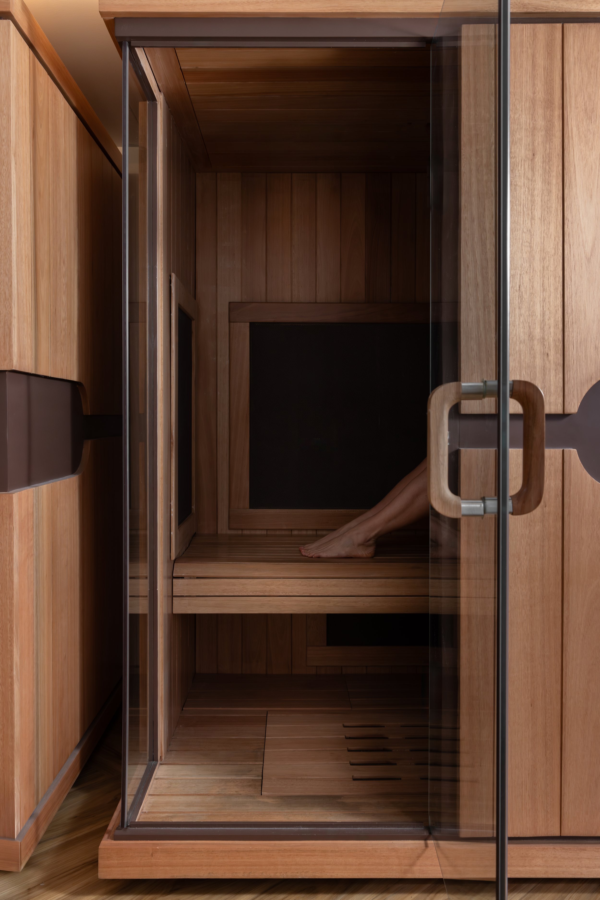 A person with light skin and bare legs sitting inside a wood-paneled sauna, visible from the knees down.