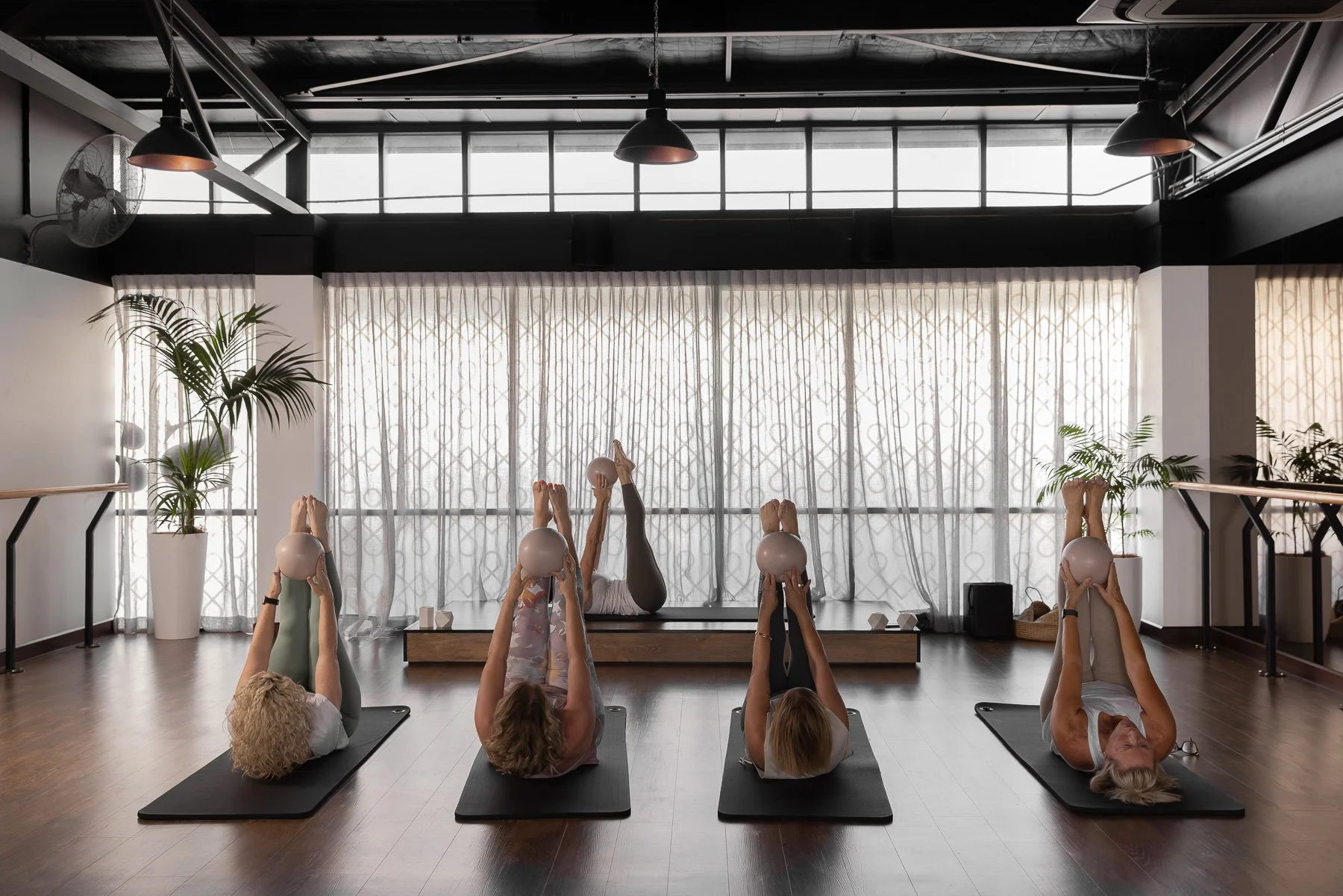 Women participating in a yoga class lying on mats, holding exercise balls above their heads, in a bright studio with large windows, plants, and industrial decor.
