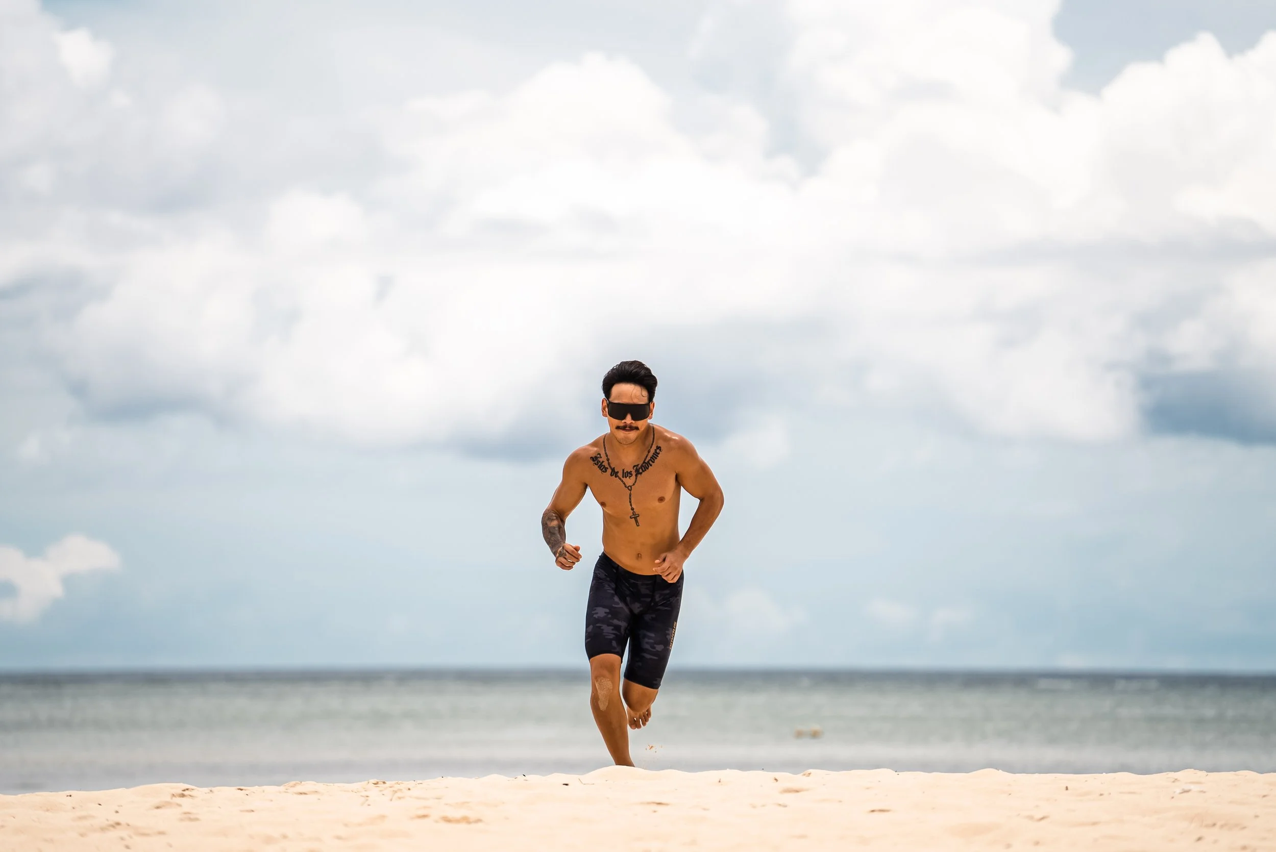 Product photography featuring a model running on a sandy beach with the ocean and cloudy sky in the background.