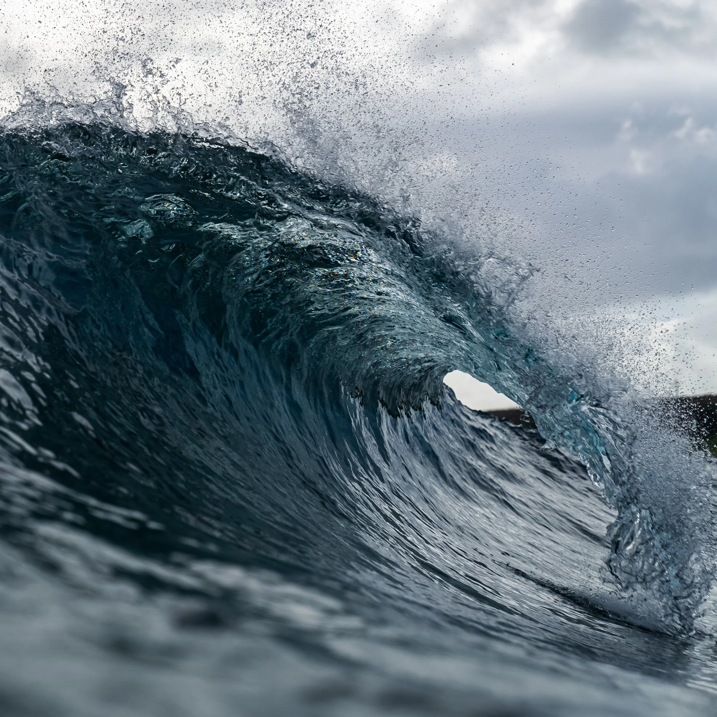 Ocean wave curling, water splashing, cloudy sky in background.