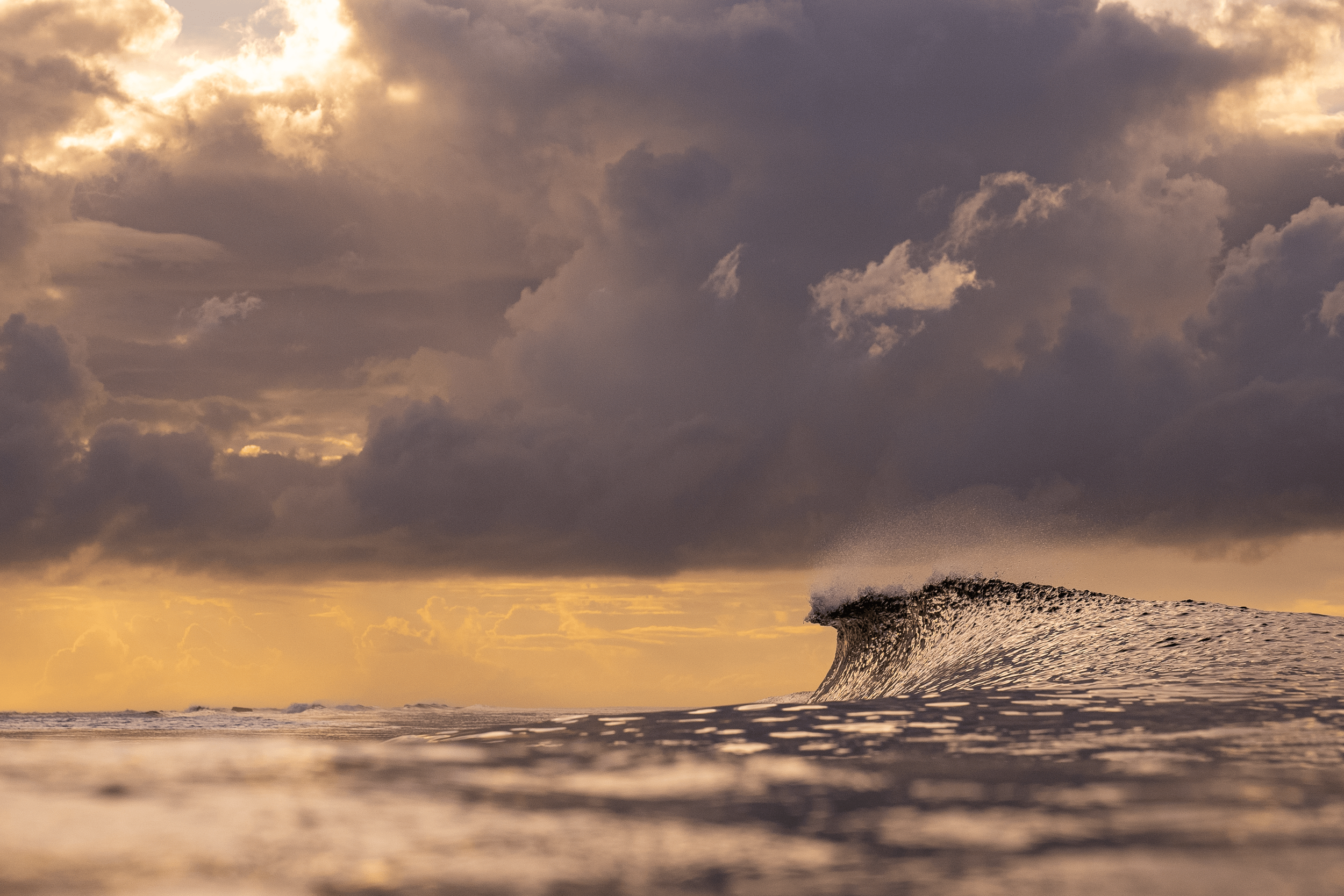 Sunset over the ocean with dark clouds and a breaking wave in the foreground.