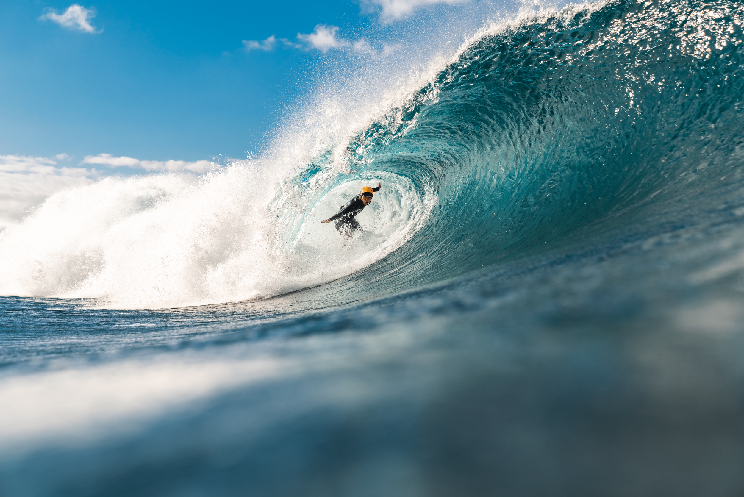 A surfer riding inside the barrel of a large blue ocean wave under a partly cloudy sky.