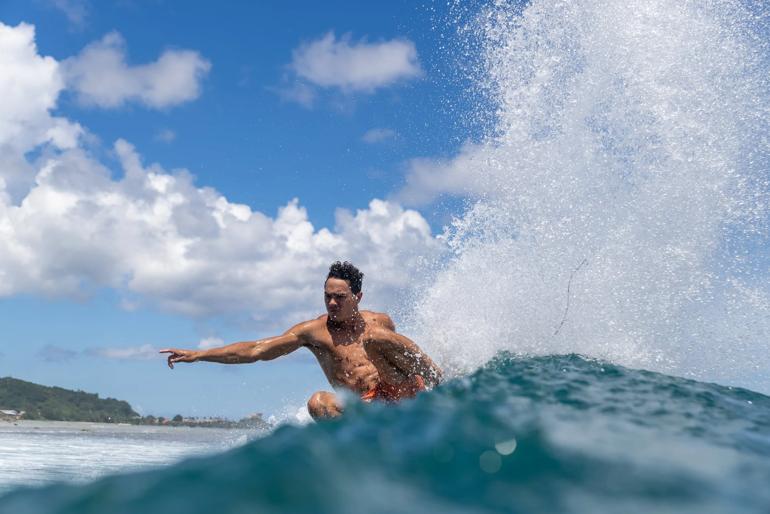 A male surfer in orange shorts riding a wave on a sunny day with a partly cloudy sky.