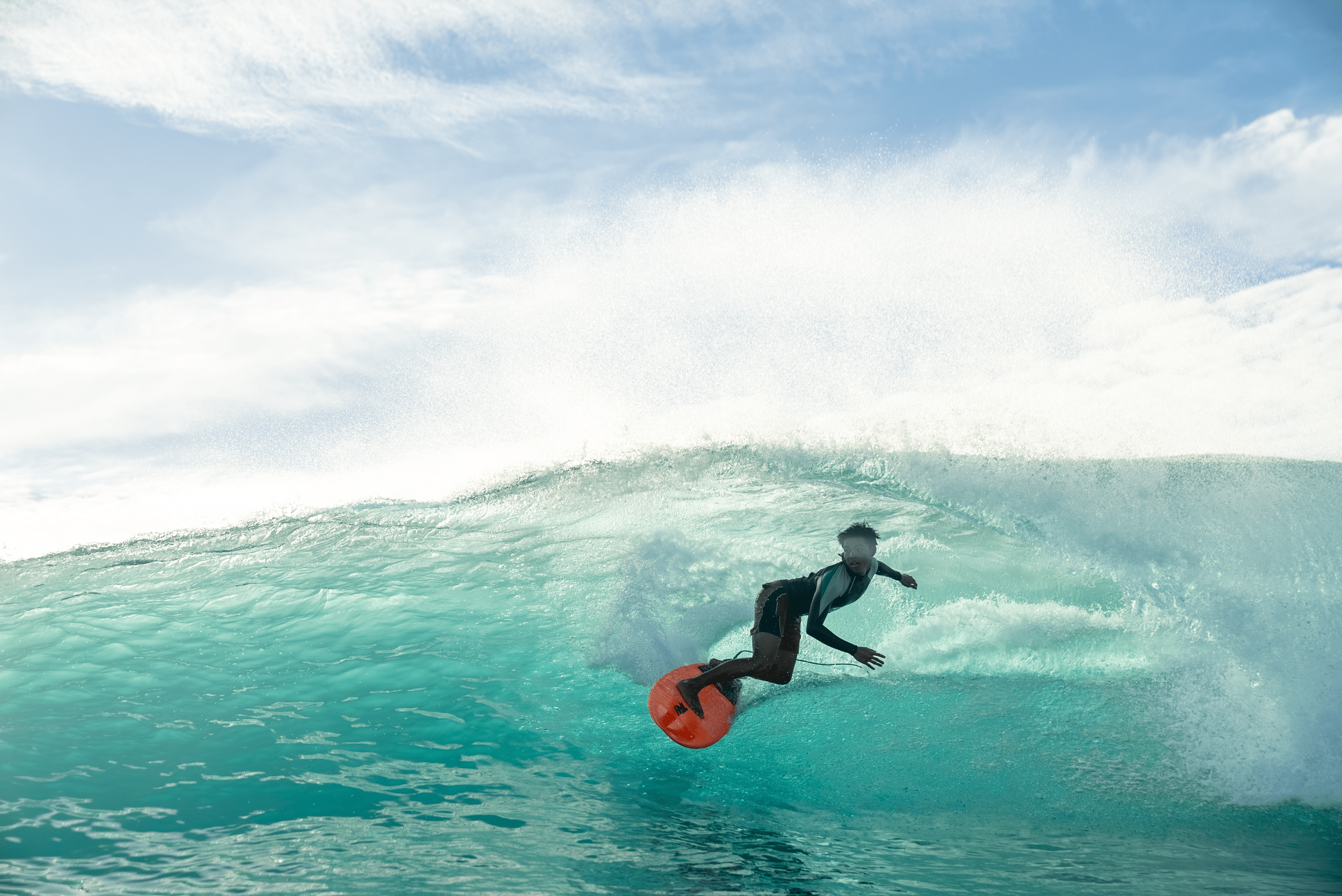 A person surfing on a wave in the ocean, with a blue sky and clouds in the background.