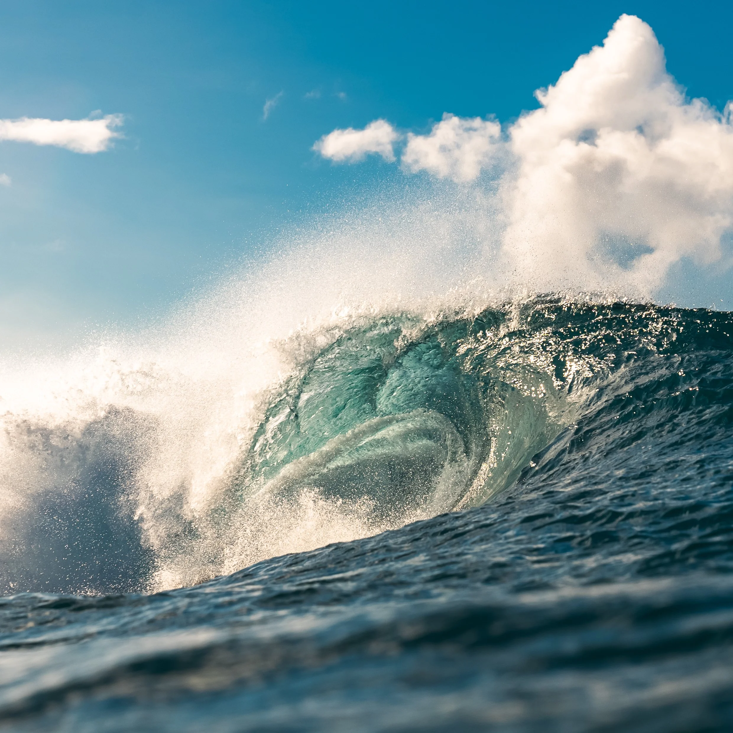 An ocean wave curling under a blue sky with clouds.