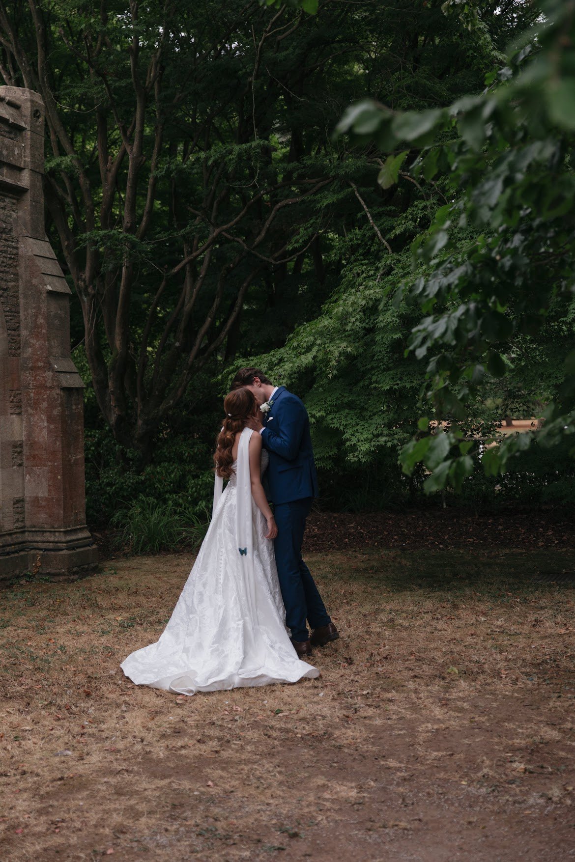 Bride and groom standing amongst trees. Bride is wearing a bridal gown and scarf with 2 blue butterflies attched