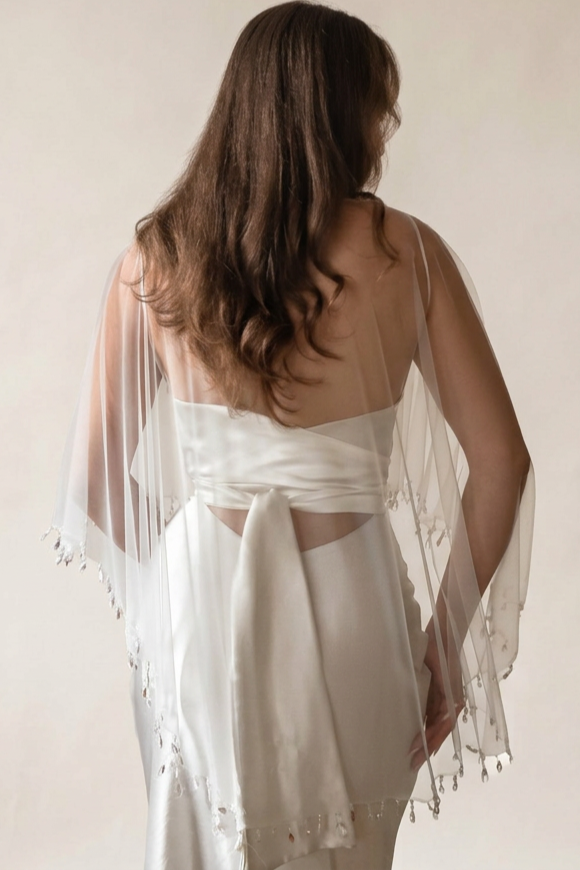 The back of a bride wearing an off-white tulle wedding cape shawl with pearl and glass crystal droplets along the edge