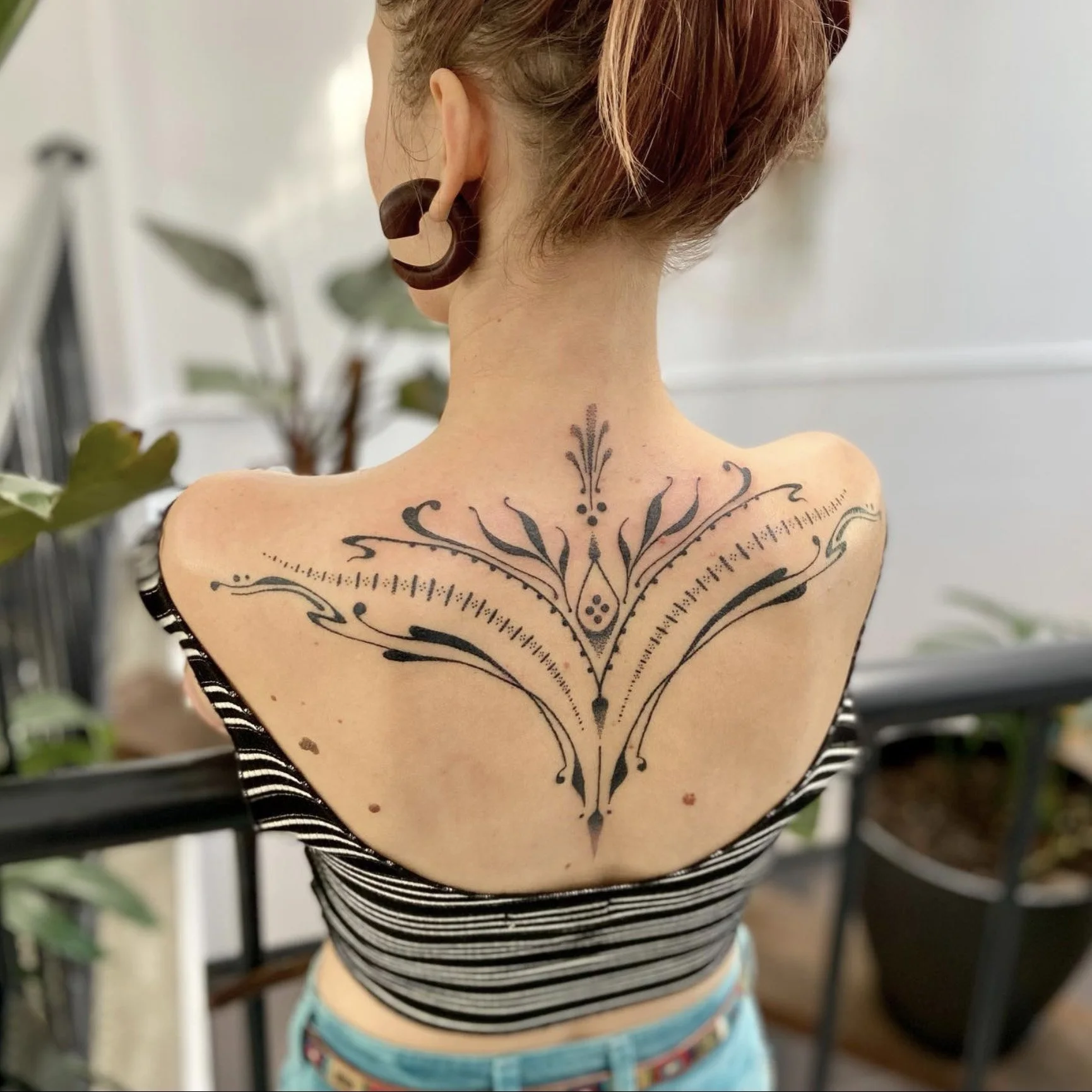 Back of a woman with a large black decorative tattoo on her upper back and a tribal-style earring.