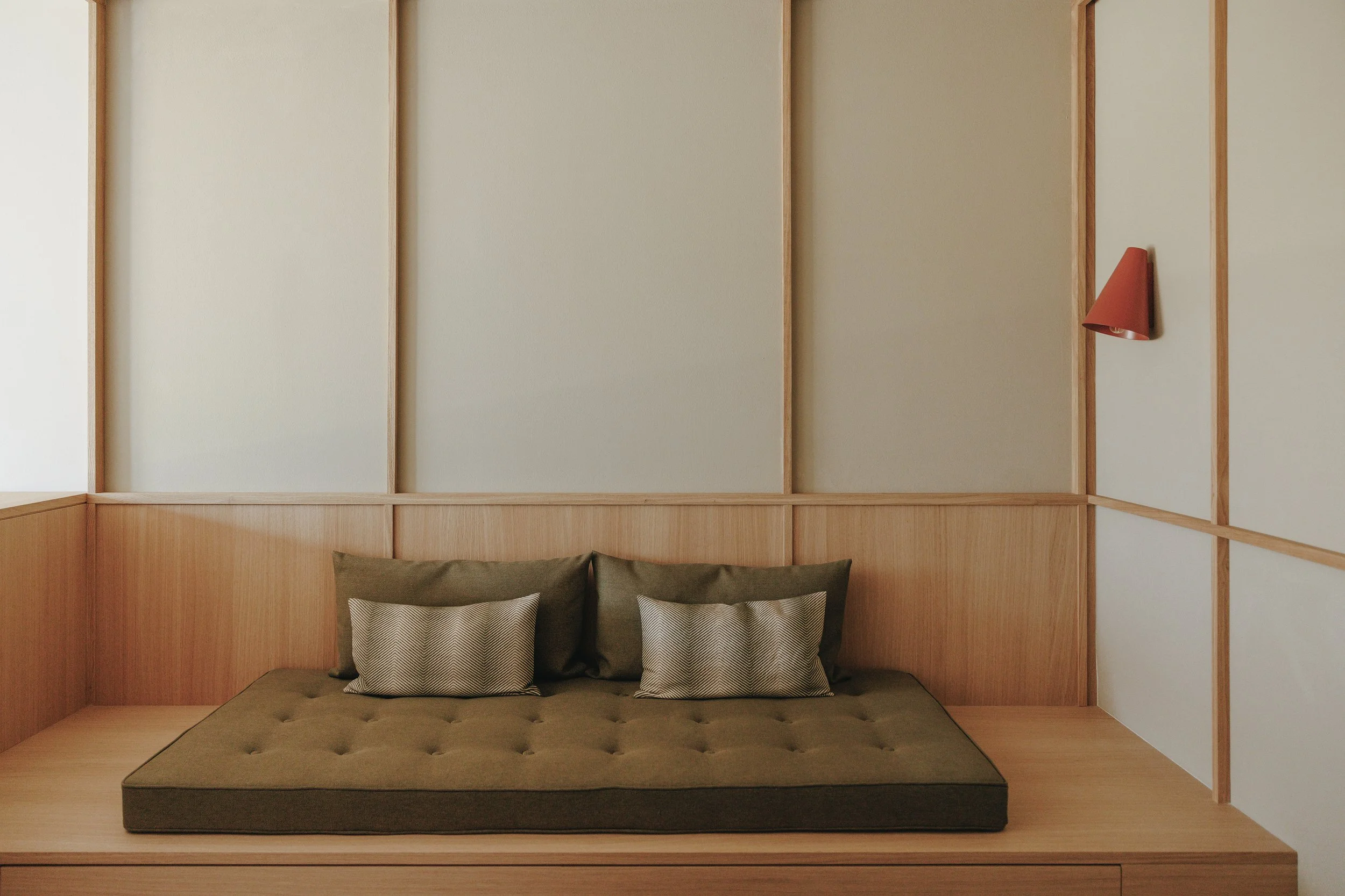 Minimalist bedroom corner with a platform bed, green mattress, two large matching pillows, and two smaller patterned throw pillows, wood paneling, and a wall-mounted red lamp.